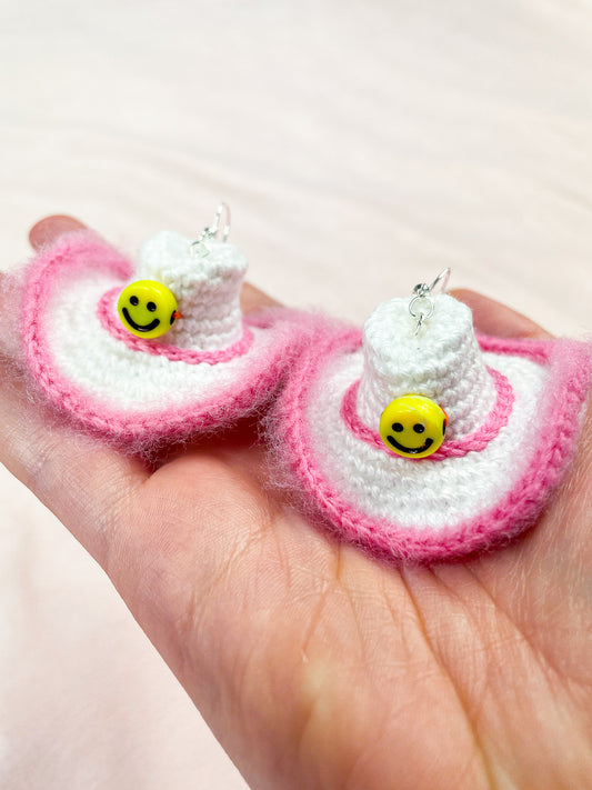 A hand holding a pair of white earrings shaped like cowboy hats with pink edges and a hatband and a yellow smiley face bead on the front.