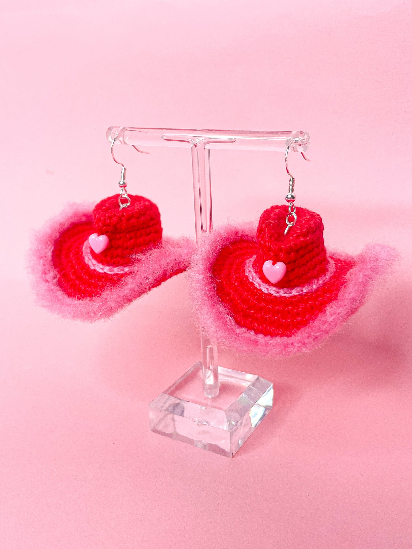 Side view of crocheted earrings shaped like hats in red with pink detailing and a pink heart bead.