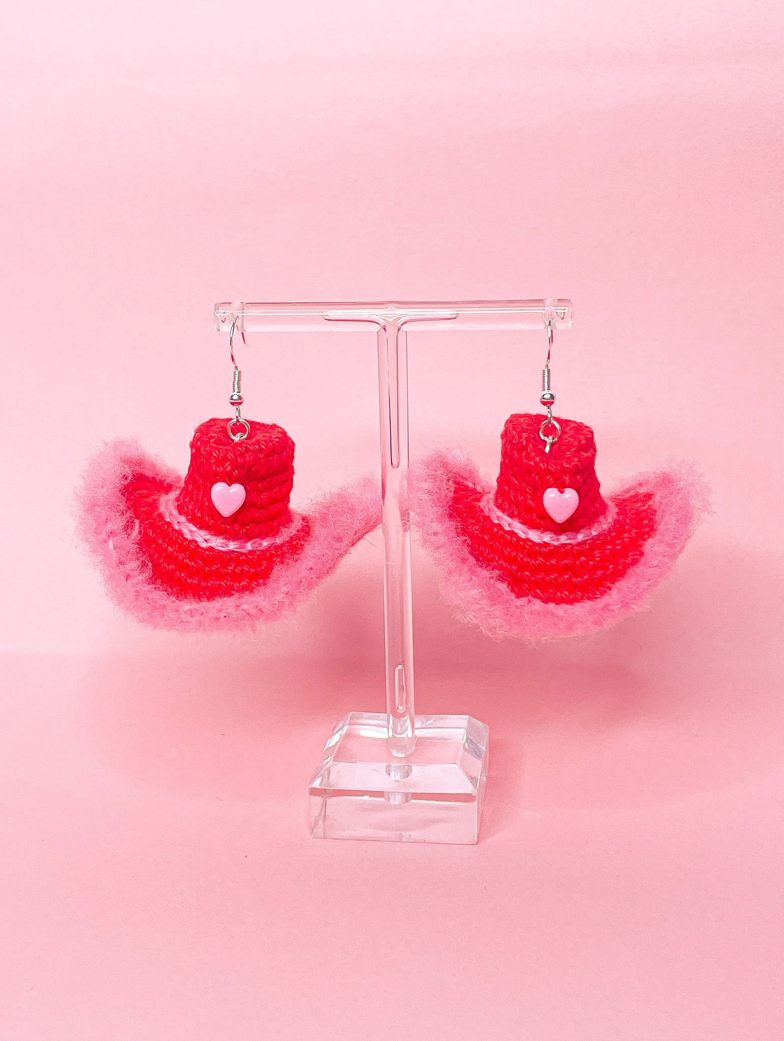 Pair of crochet cowboy hat earrings in red and pink hanging on an earring stand on a pink background.