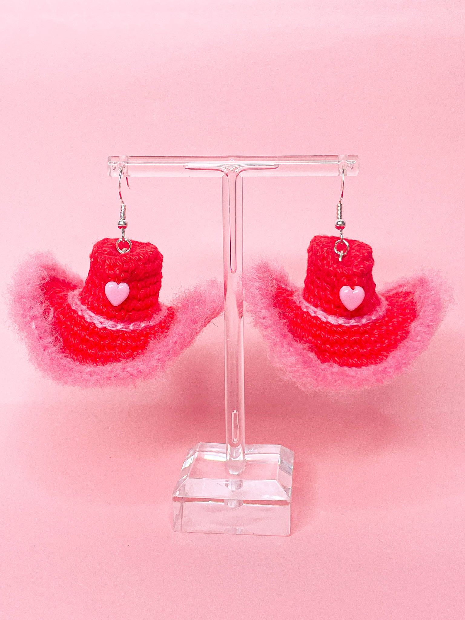 Close view of a pair of cowboy hat earrings in red with a pink fluffy edge, hatband and heart bead on the front.