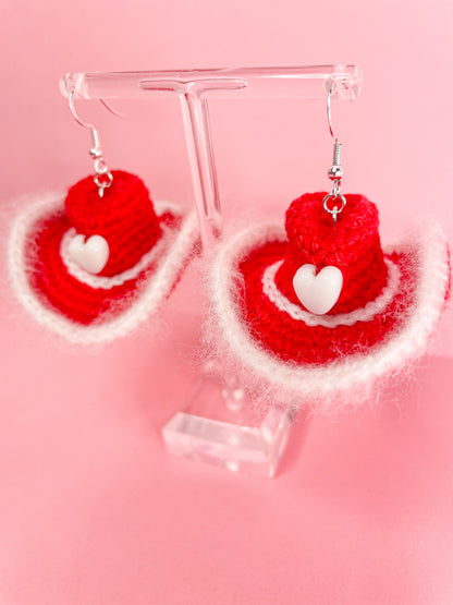 Above view of a pair of crochet cowboy hat earrings in red with white detailing on an earring stand.