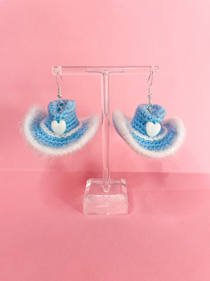 A pair of crochet earrings shaped like cowboy hat earrings in baby blue hanging on an earring stand.