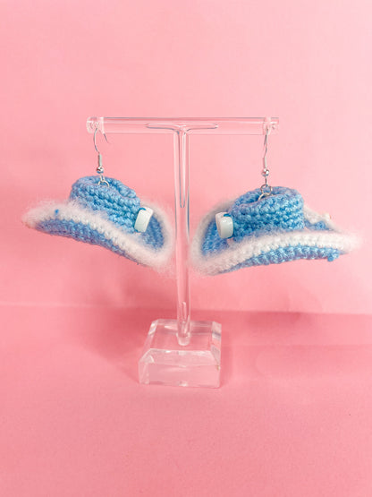 Side view of a pair of baby blue crochet cowboy hat earrings hanging on an earring stand.