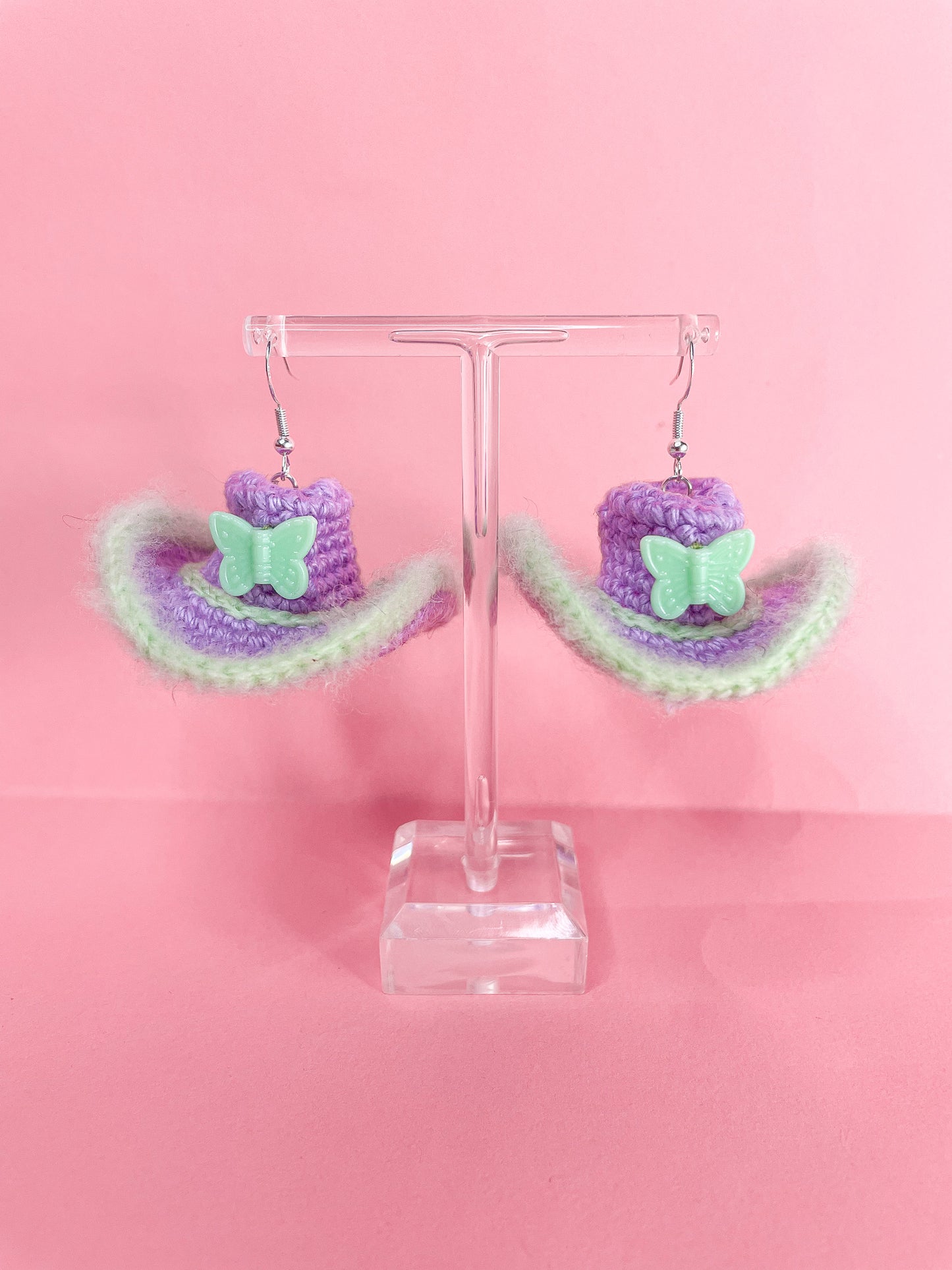 Pair of cowboy hat shaped earrings in purple and green hanging on an earring stand.