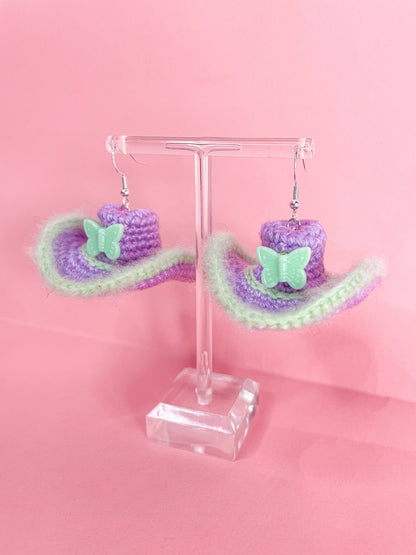 Above view of a pair of lilac crochet earrings resembling cowboy hats with green edges handing on an earring stand.