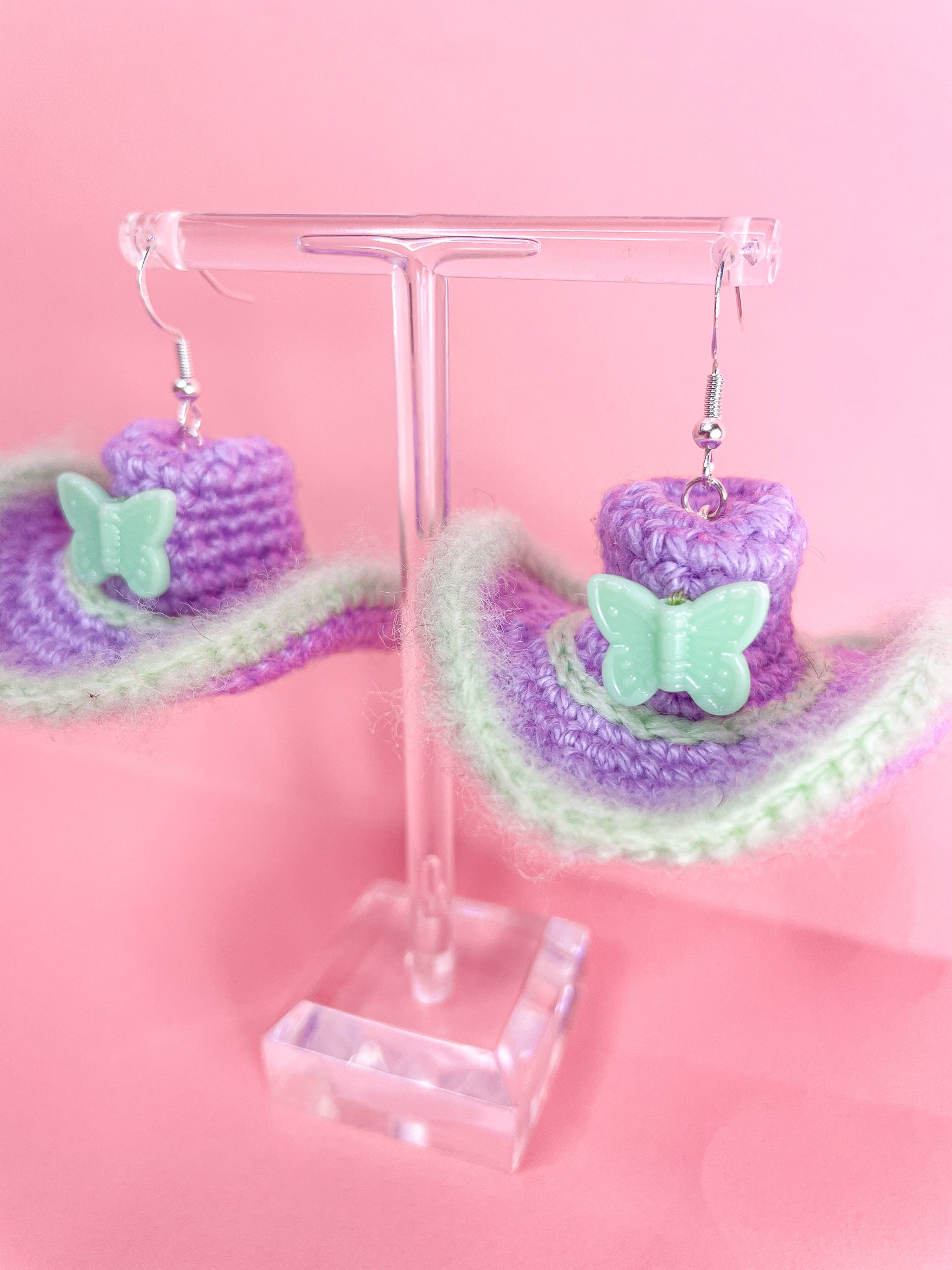 Close up of a pair of lilac and green crochet earrings shaped like cowboy hats on an earring stand.