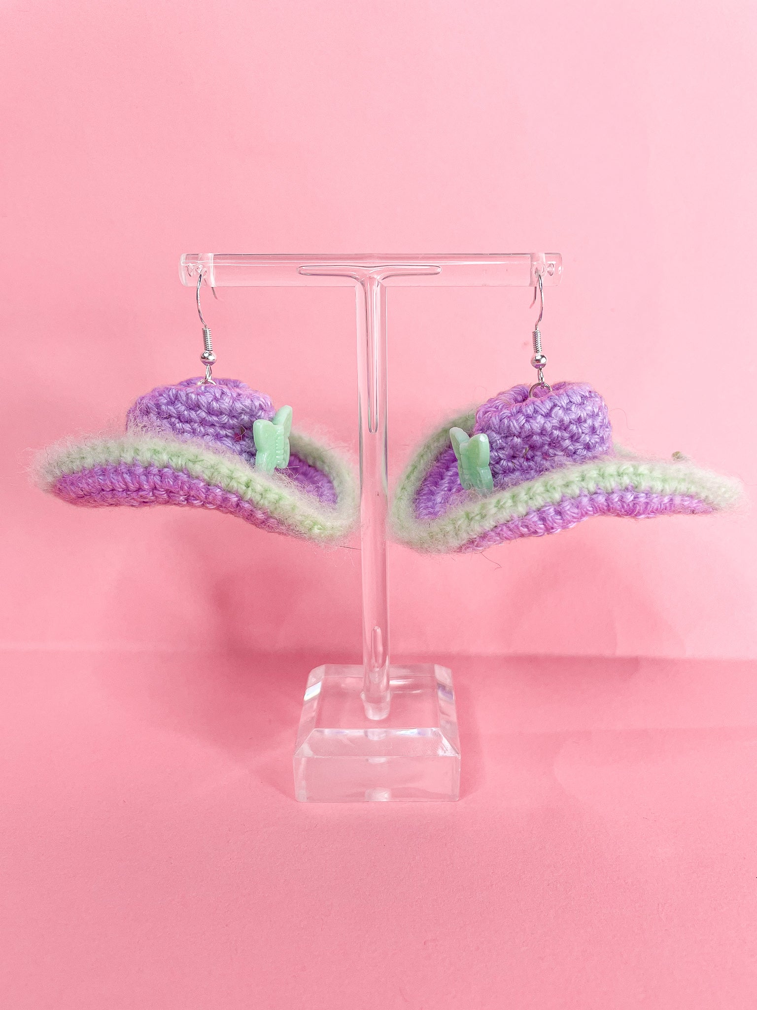 Side view of a pair of crochet cowboy hat earrings in purple and green hanging on an earrings stand.