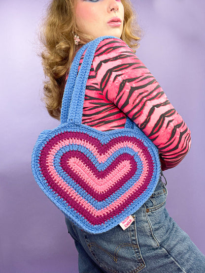 Side view of woman wearing a crochet heart bag resembling Bisexual flag colours (pink, purple, blue) with a matching outfit.