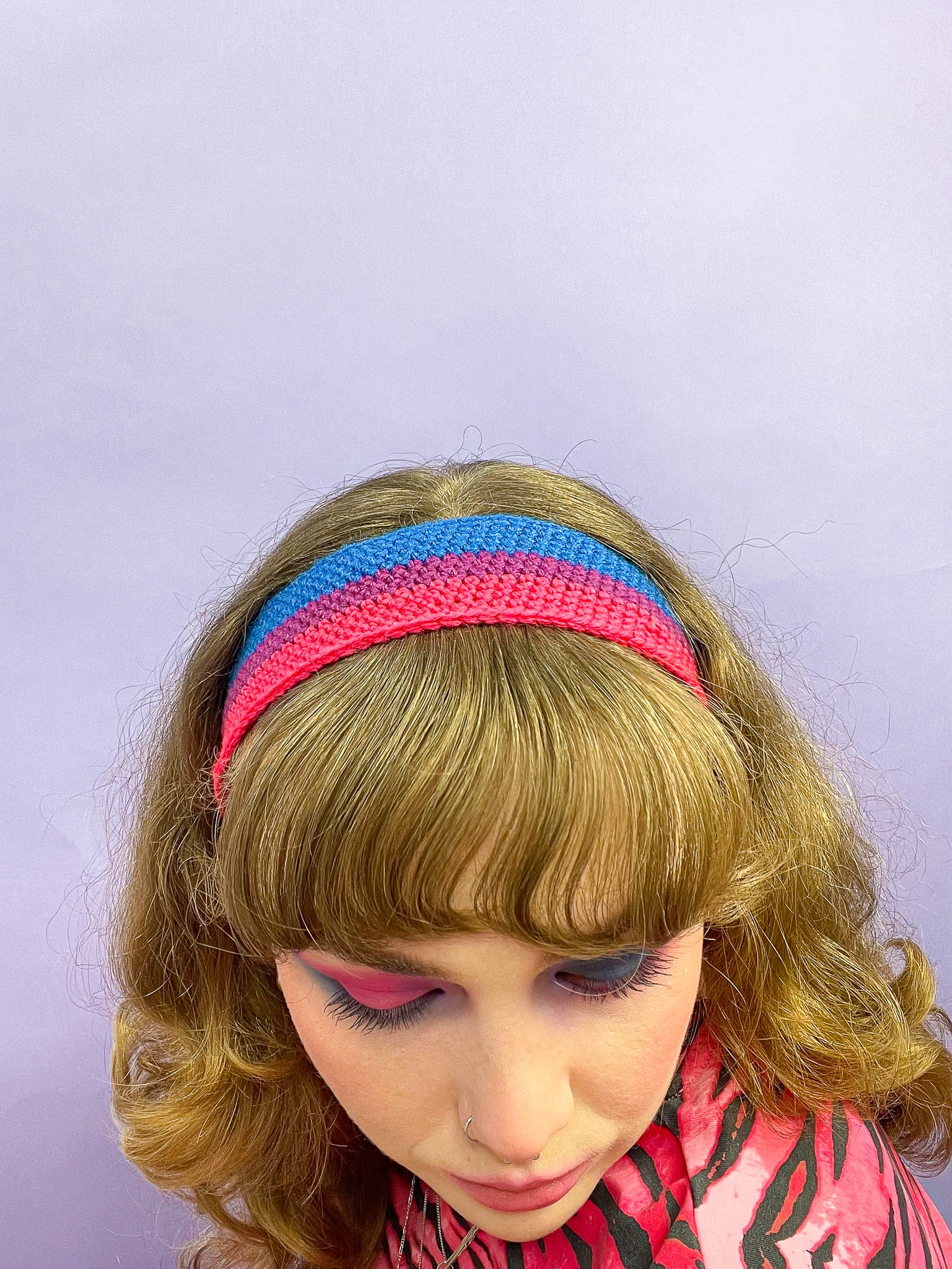 Above view of a woman wearing a crocheted headband resembling the bisexual flag colours (pink, purple and blue) with a matching top and makeup.