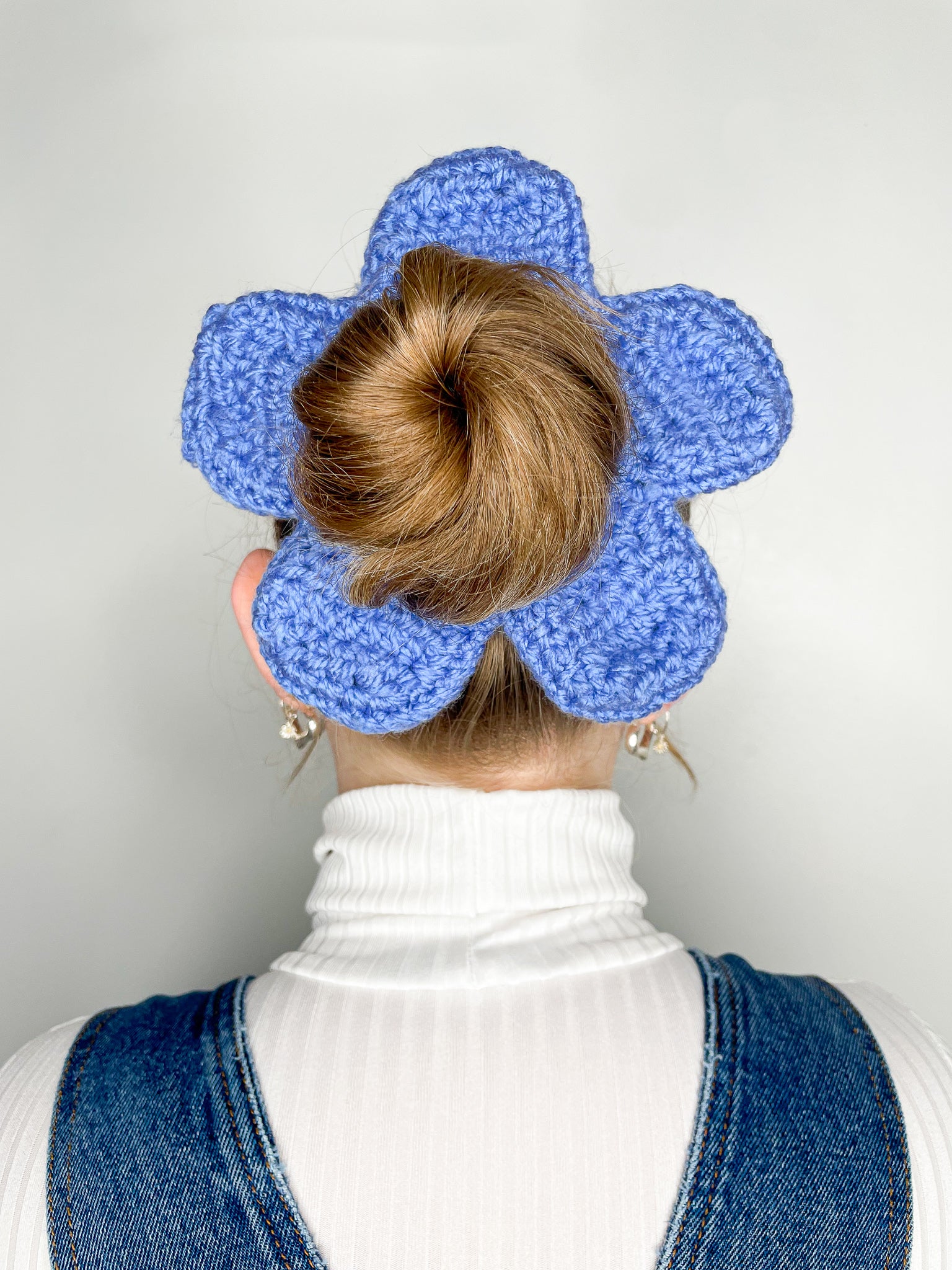 Back view of a woman wearing a blue crochet scrunchie shaped like a daisy in her hair with a white turtleneck and denim dungarees.