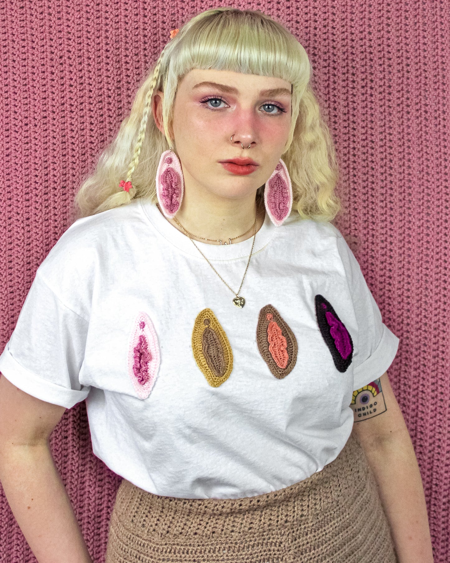 A woman wearing a white t-shirt with four crocheted vulvas sewn on the front and matching earrings, paired with brown crochet pants.