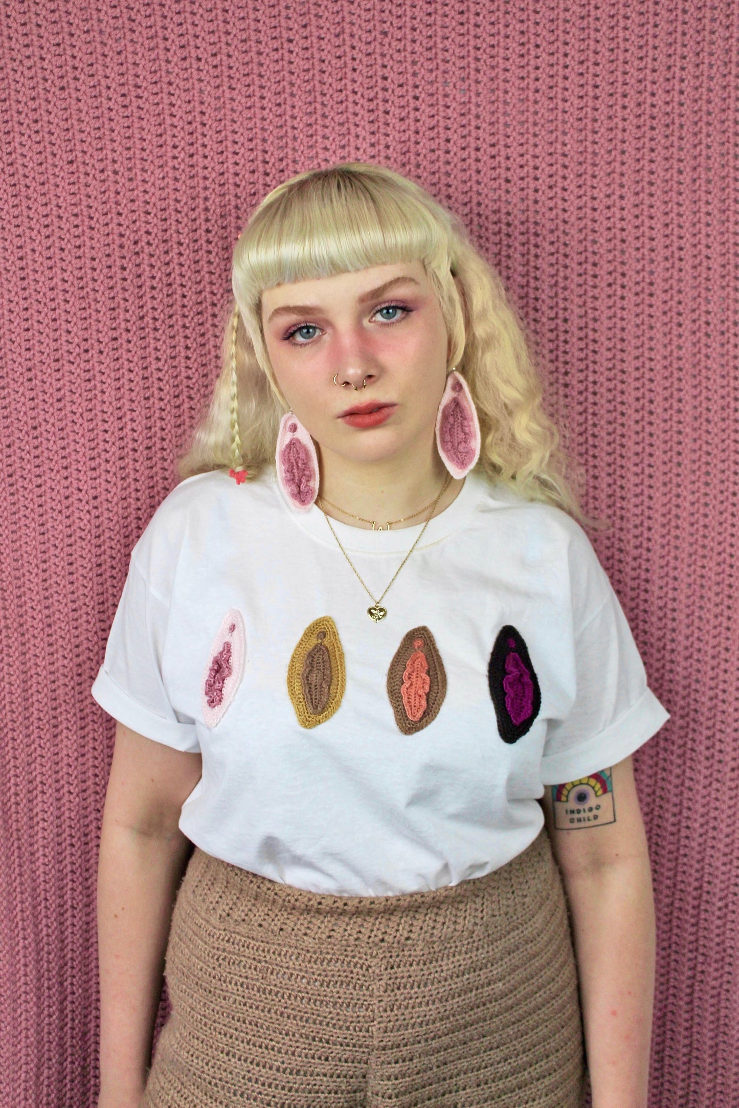 A woman wearing a matching pair of crochet vulva earrings and appliquéd t shirt in white with various vulva shades on the front.