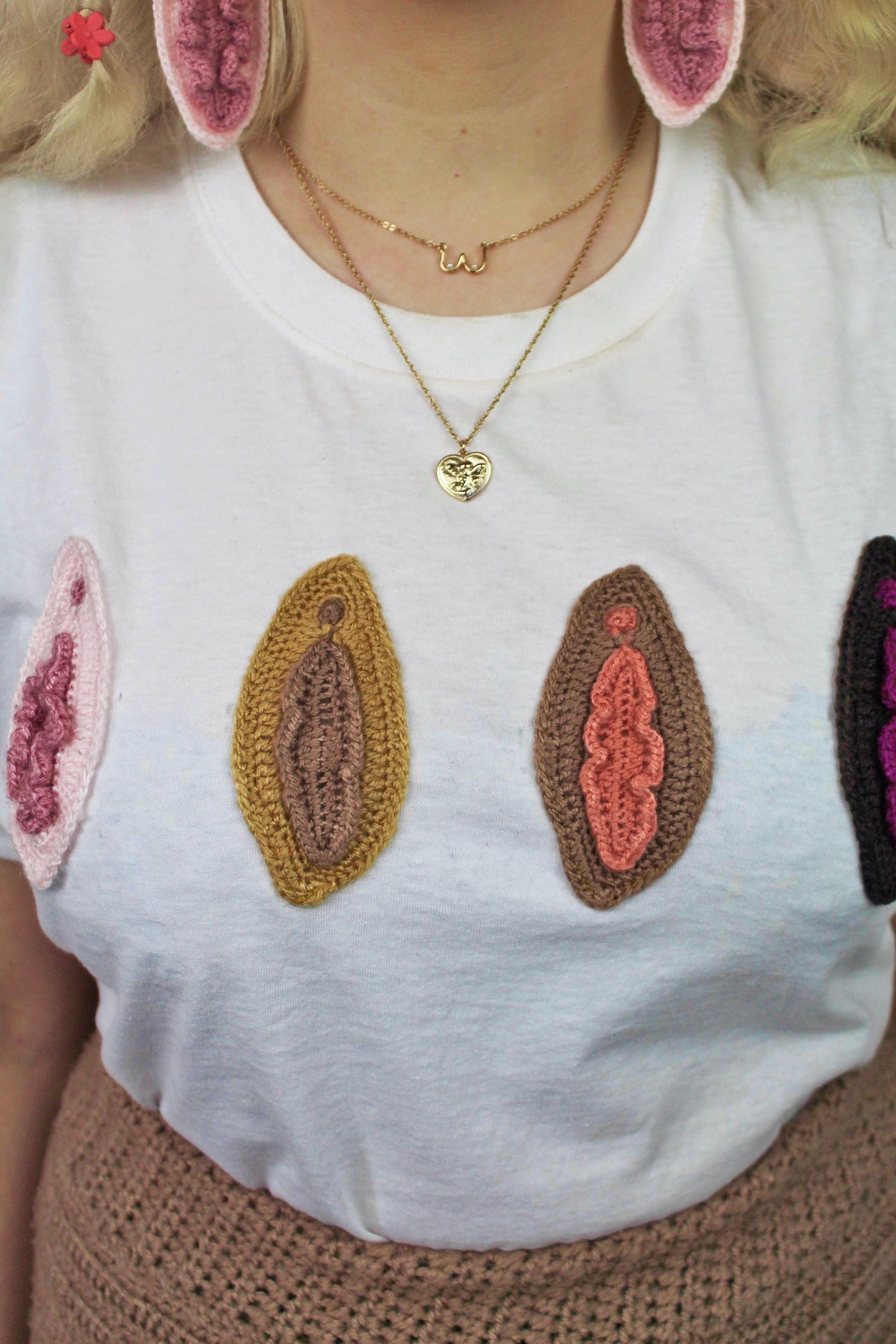 Close up of a woman wearing a white t shirt with different shades of appliqué vulvas attached to the front and a matching pair of earrings and crochet pants.