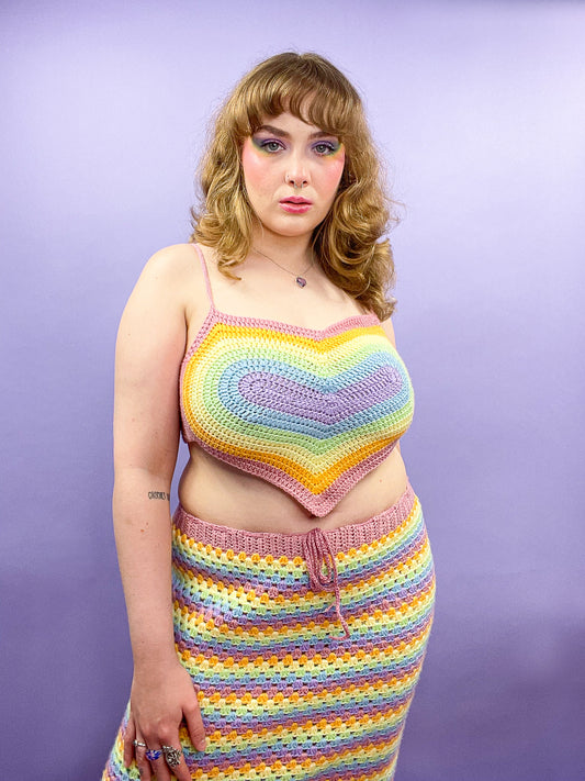 A woman posing wearing  a heart shaped crochet top in pastel rainbow colours with a matching striped skirt standing in front of a purple background.