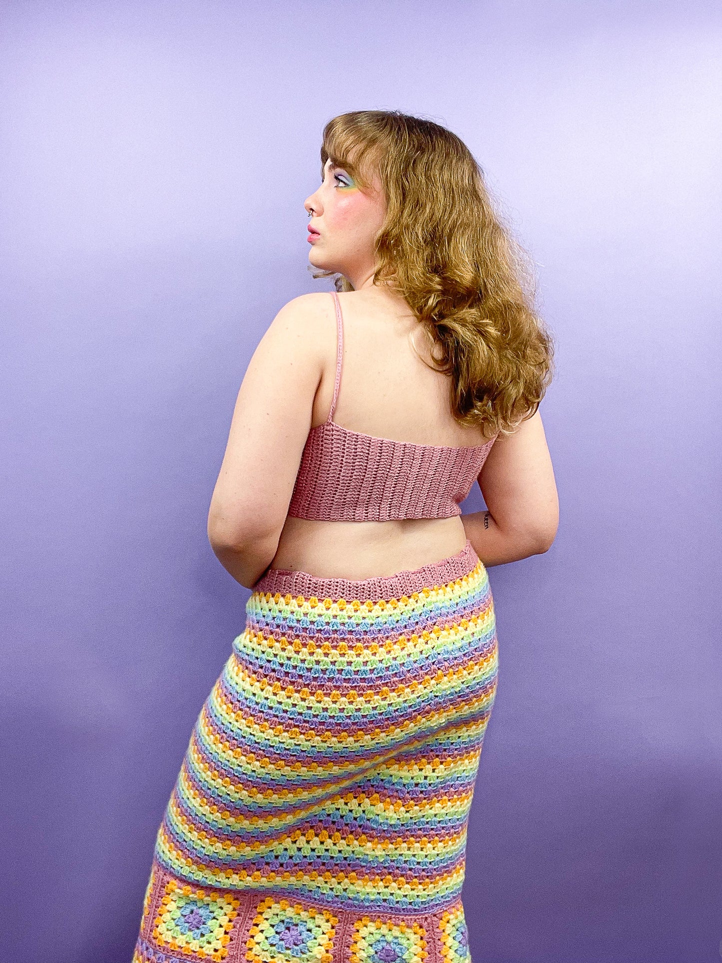 Back view of a woman posing wearing a crochet rainbow pastel skirt with a pale pink crop top.