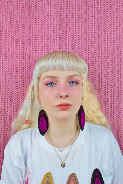 Close view of a woman's facing wearing a pair of statement earrings shaped like vaginas in dark brown with a matching t shirt that has crochet appliqué vulvas.