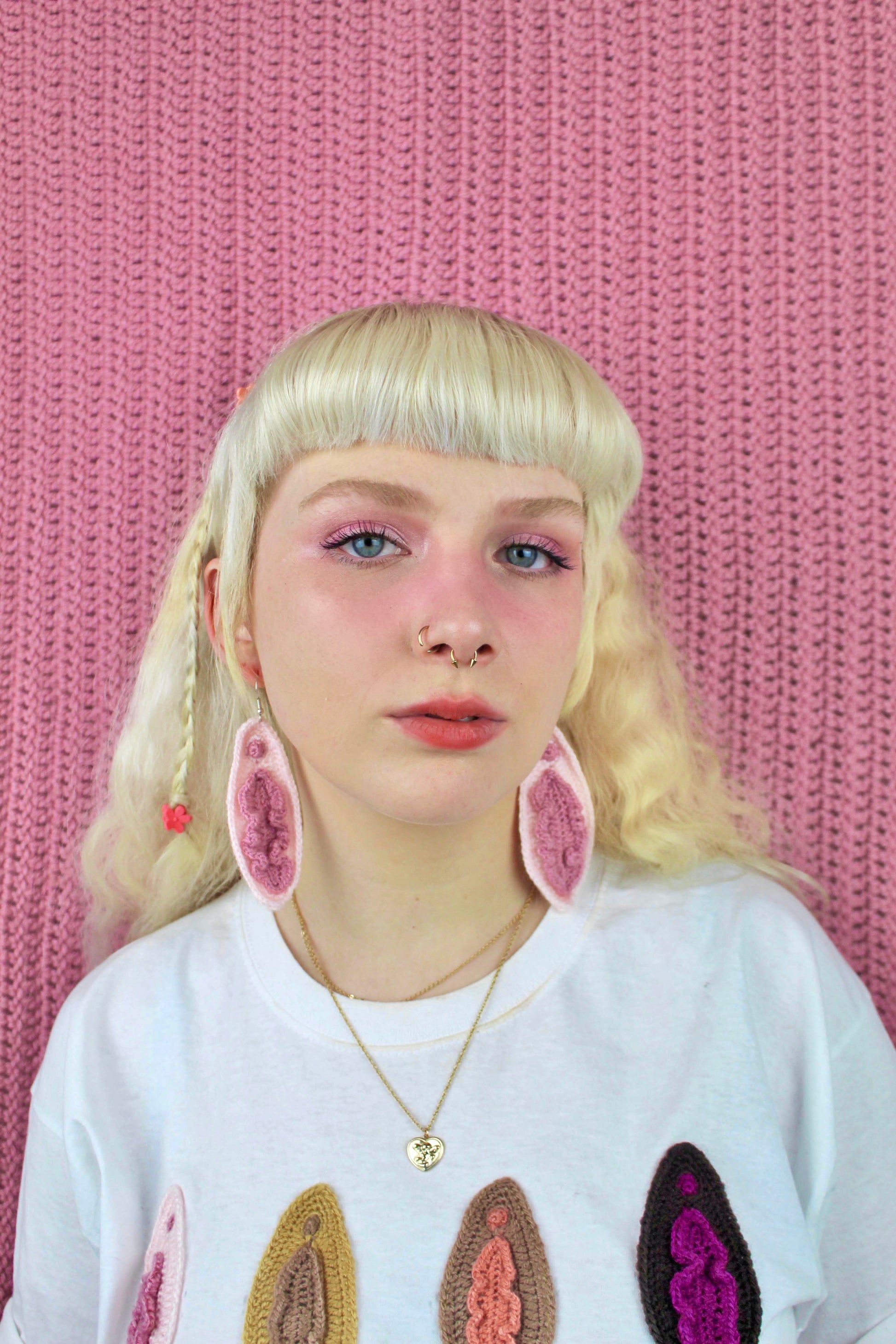 Close up of a woman wearing a pair of vulva inspired earrings in cream with pink details and a matching t shirt with appliqué vulvas on the front.