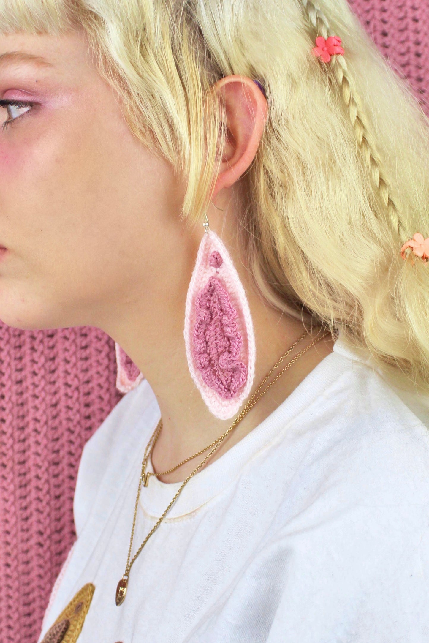 Side view of a woman wearing a pair of vulva earrings in cream in front of a pink crochet background.