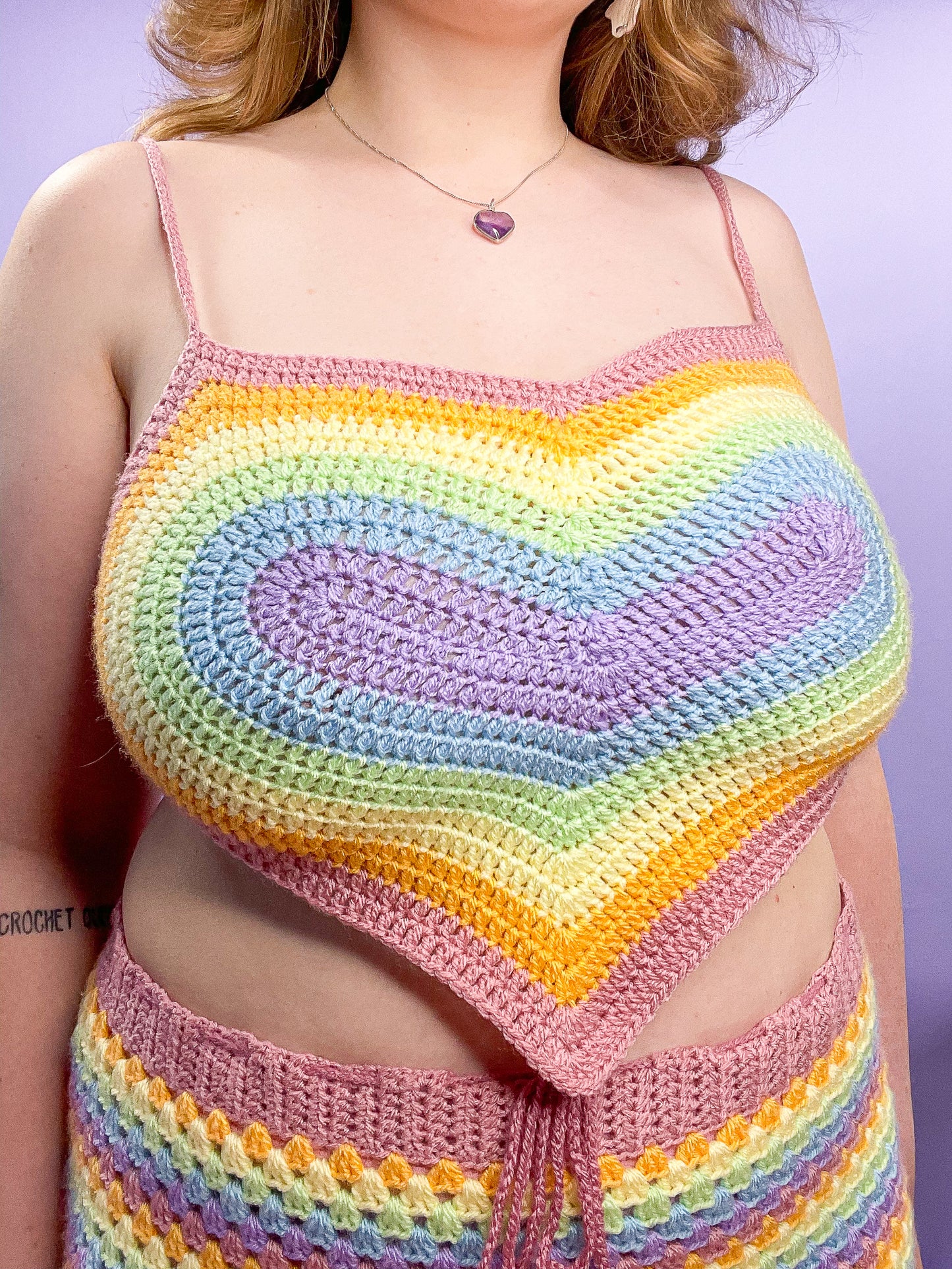 Close up of a woman wearing a crochet heart top in pastel rainbow colours with a matching skirt and lilac heart pendant.