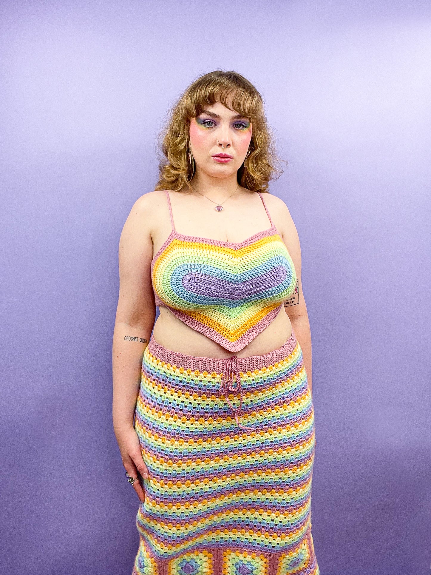 Front view of a woman standing wearing a pastel rainbow crochet set with a heart shaped top and striped skirt.