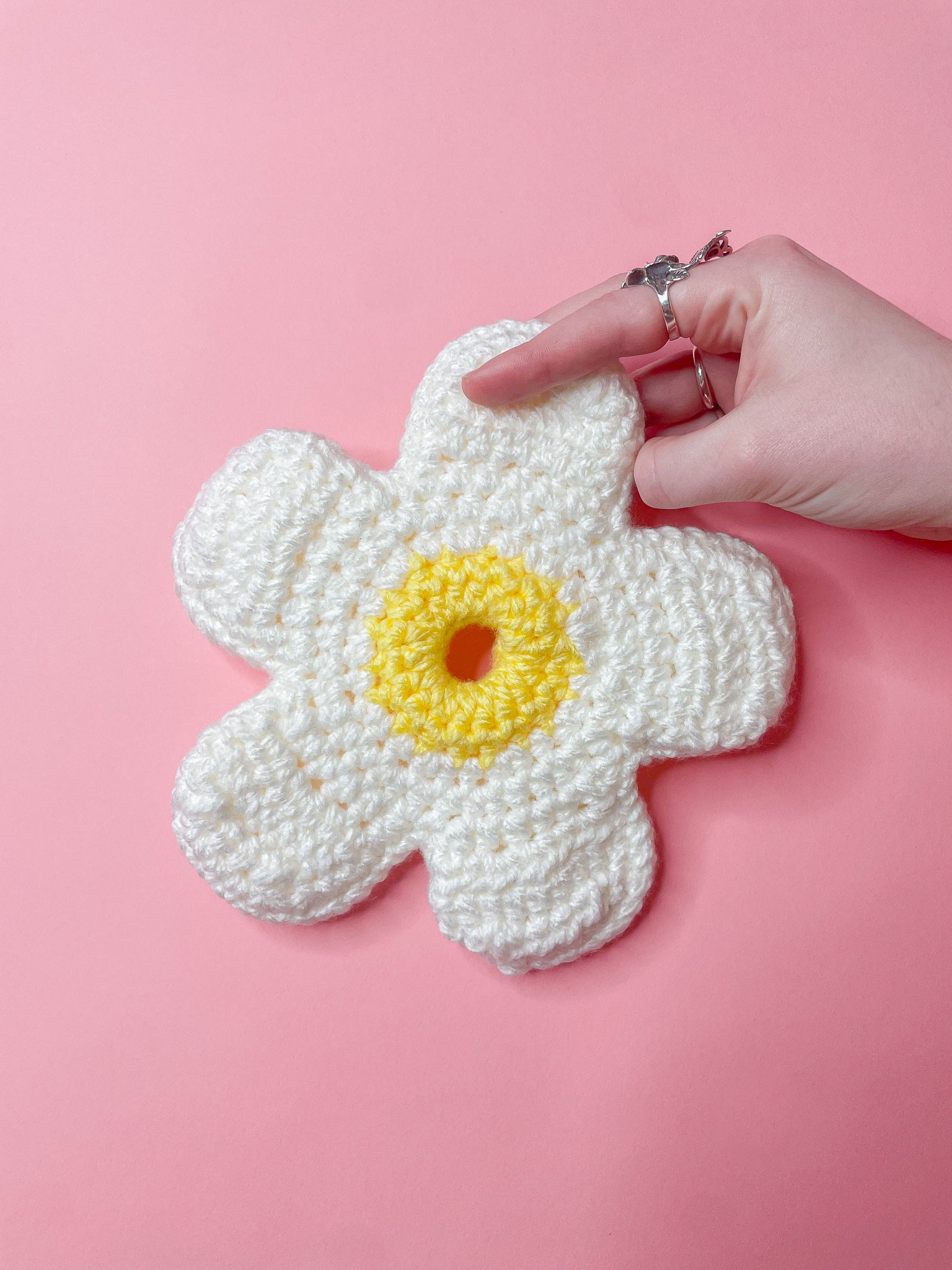 A hand holding a white daisy scrunchie with a yellow centre against a pink background.