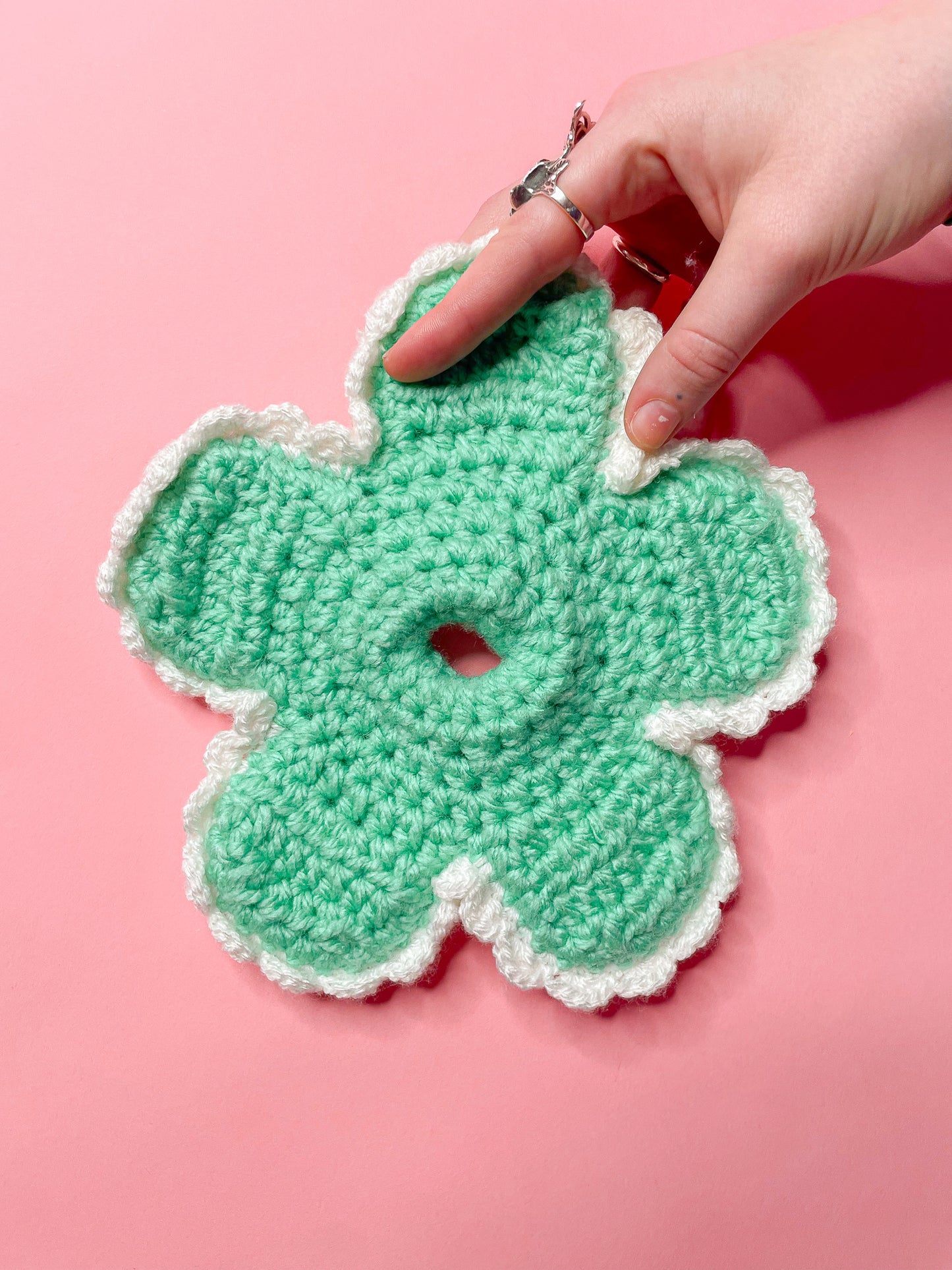 A hand holding a daisy crochet scrunchie in green with white frills against a pink background.