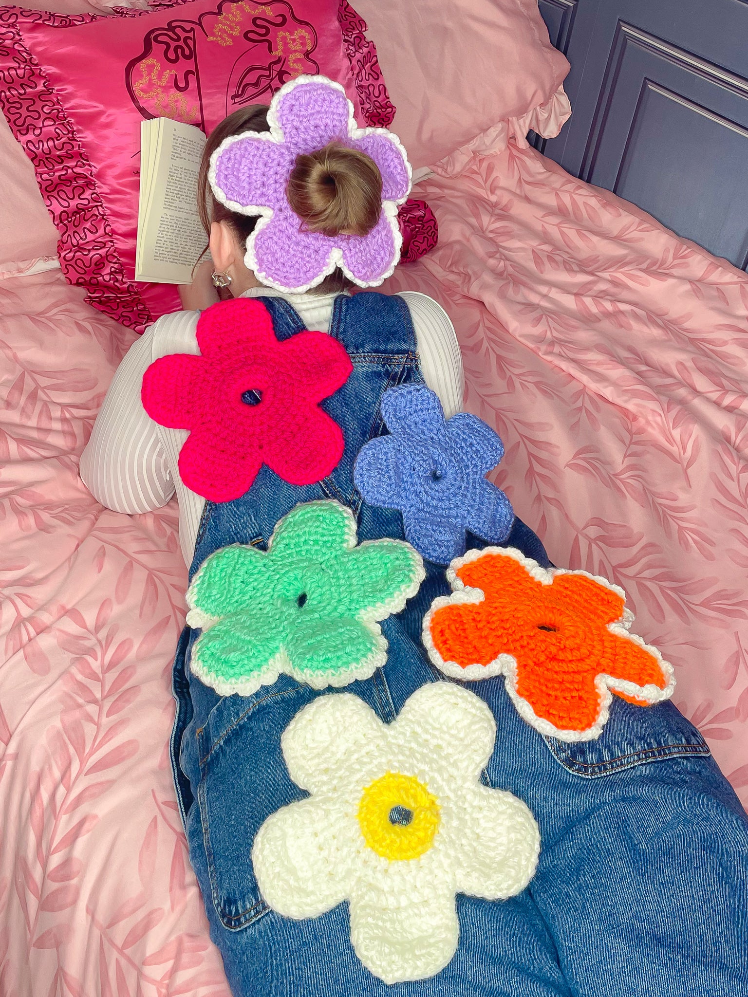 Back view of a woman lying on a bed reading a book wearing a purple flower shaped scrunchie in her hair and five scrunchies lying on her back in different colours.