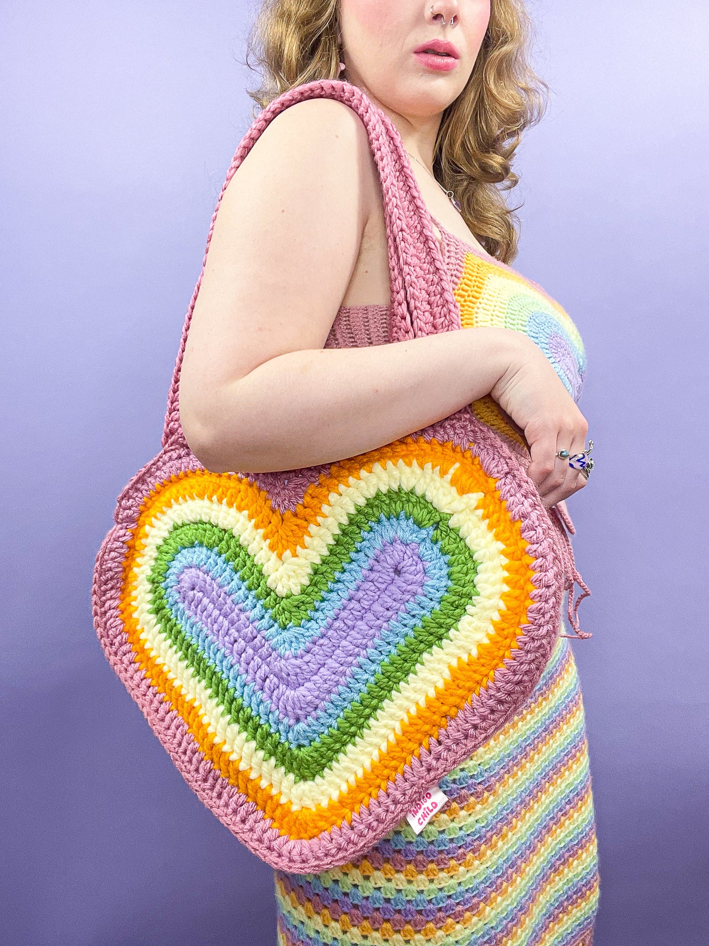 Side view of a woman wearing a crochet bag shaped like a heart in pastel rainbow colours with a matching top and skirt.