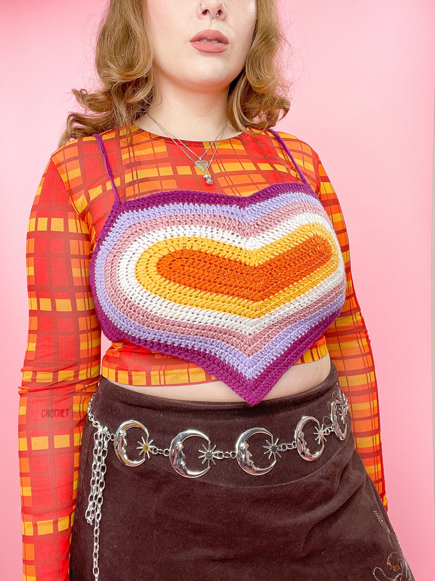 Close view of a woman wearing a crochet crop top shaped like a heart with an orange, white, pink and purple rainbow design and a matching outfit.