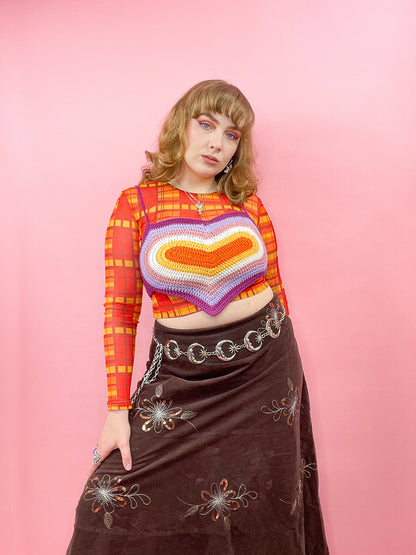 A woman posing wearing a crochet heart top in the lesbian flag colours paired with an orange gingham mesh long sleeved top, a brown cord maxi skirt and matching eyeshadow.