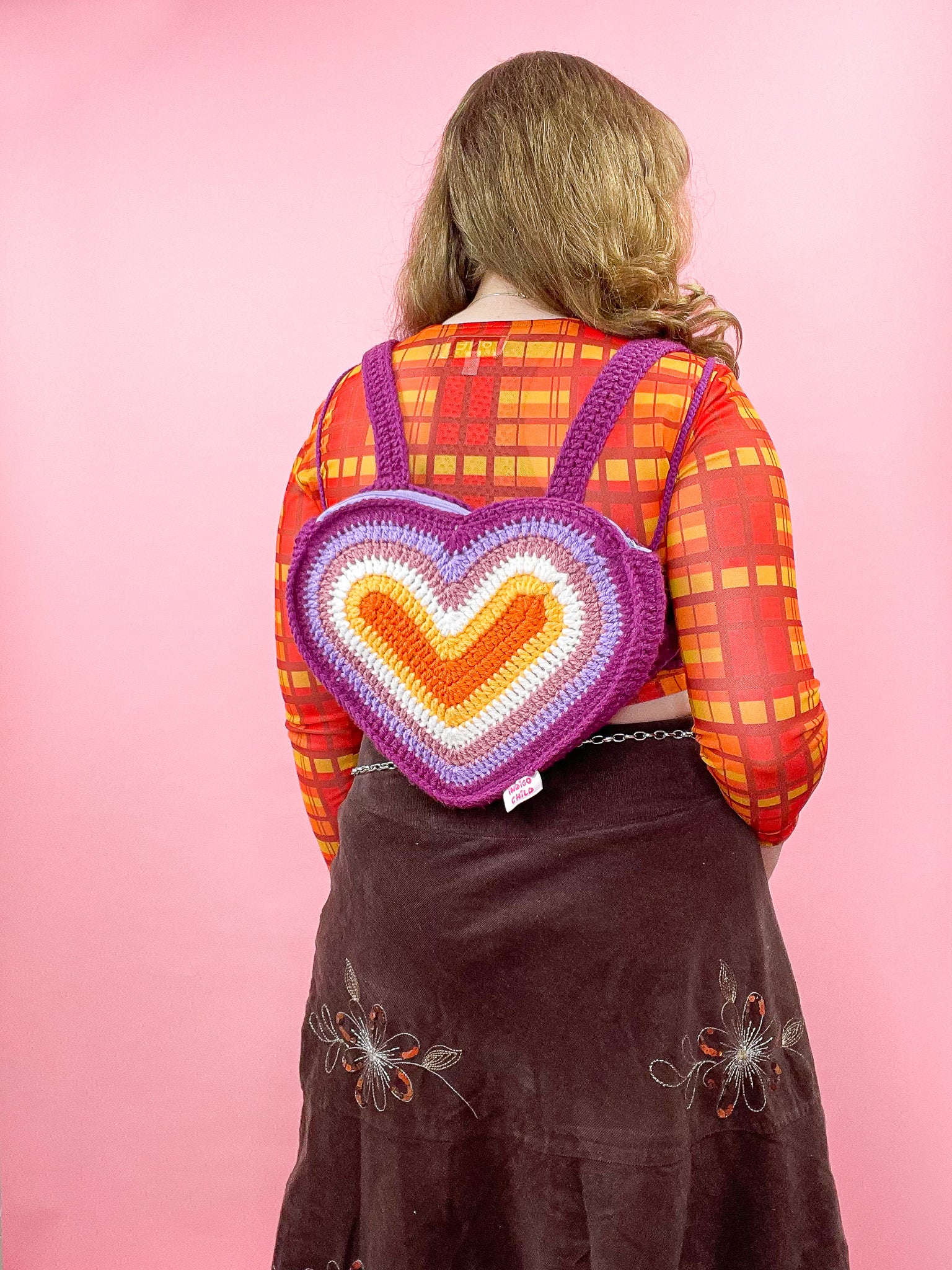 Back view of woman wearing a heart shaped backpack in a rainbow design resembling the lesbian flag colours (orange, white, pink and purple) with a matching orange top and brown skirt.