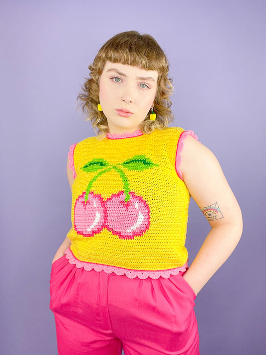 A woman wearing a yellow crochet vest with a pink cherry on the front and pink frilly edges paired with pink trousers and bell pepper earrings.