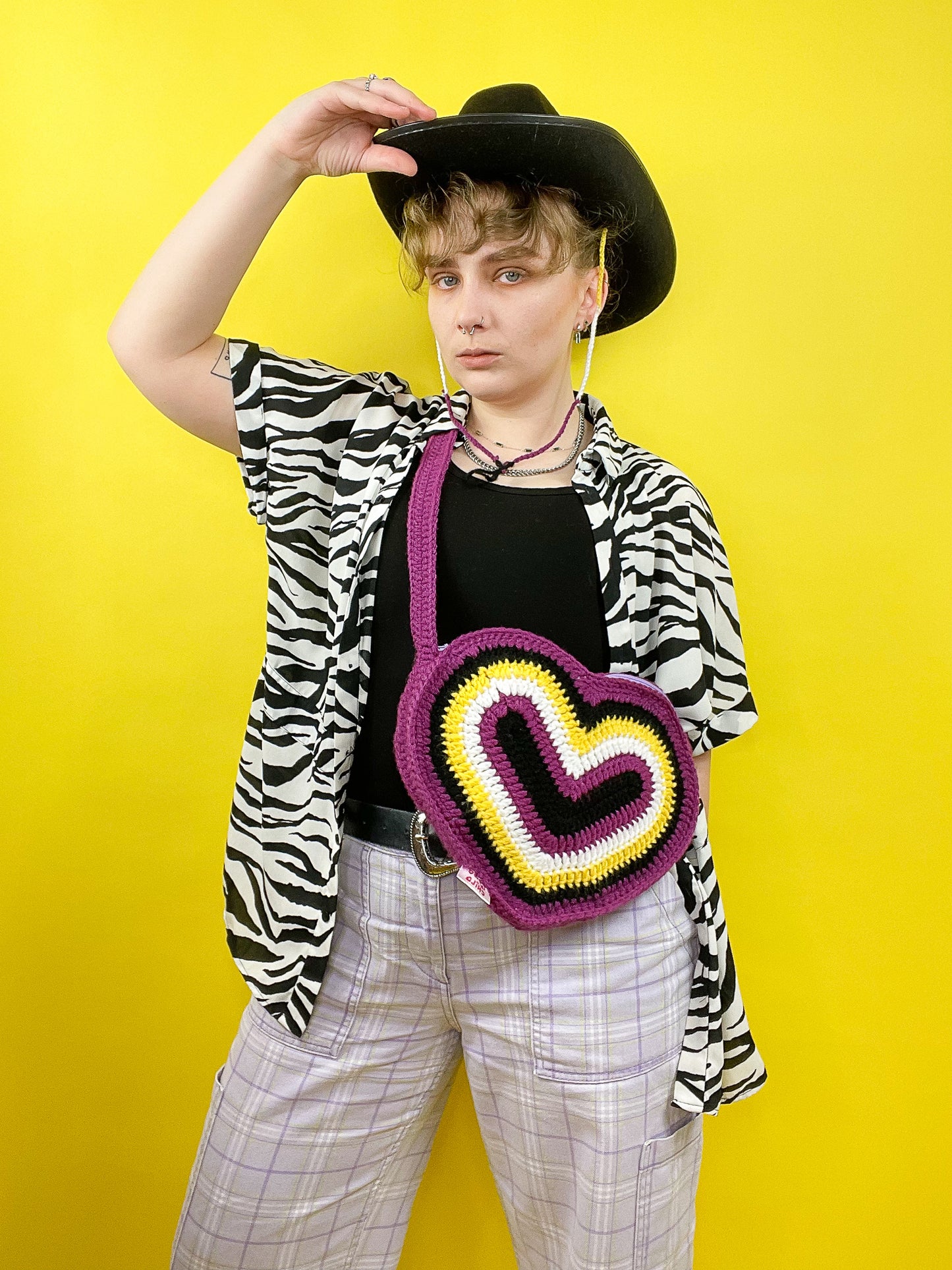 A person wearing a heart shaped crossbody bag resembling non-binary flag colours (black, purple, white, yellow) wearing a matching black binder, zebra shirt, lilac jeans and a black cowboy hat.