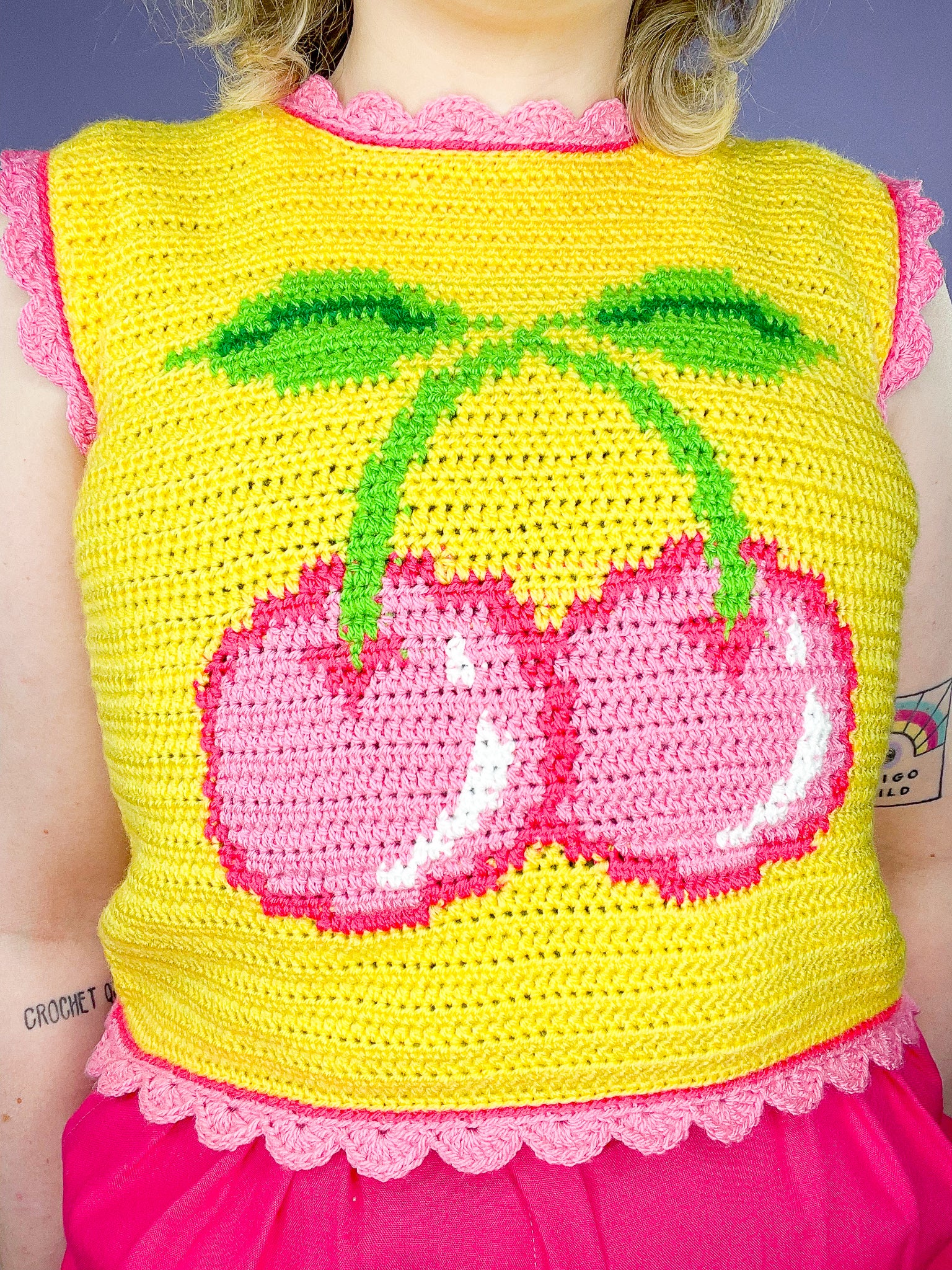 Close up of a crochet tapestry vest top in yellow with a pink cherry on the front and pink scalloped edges on the hemlines.