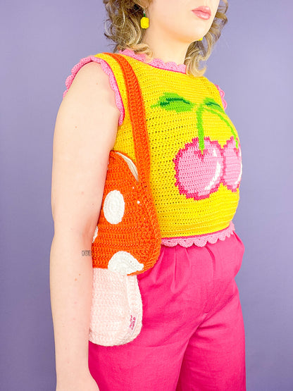 Side view of a woman wearing an orange mushroom shaped bag with a yellow vest that has a pink cherry on the front paired with pink trousers.