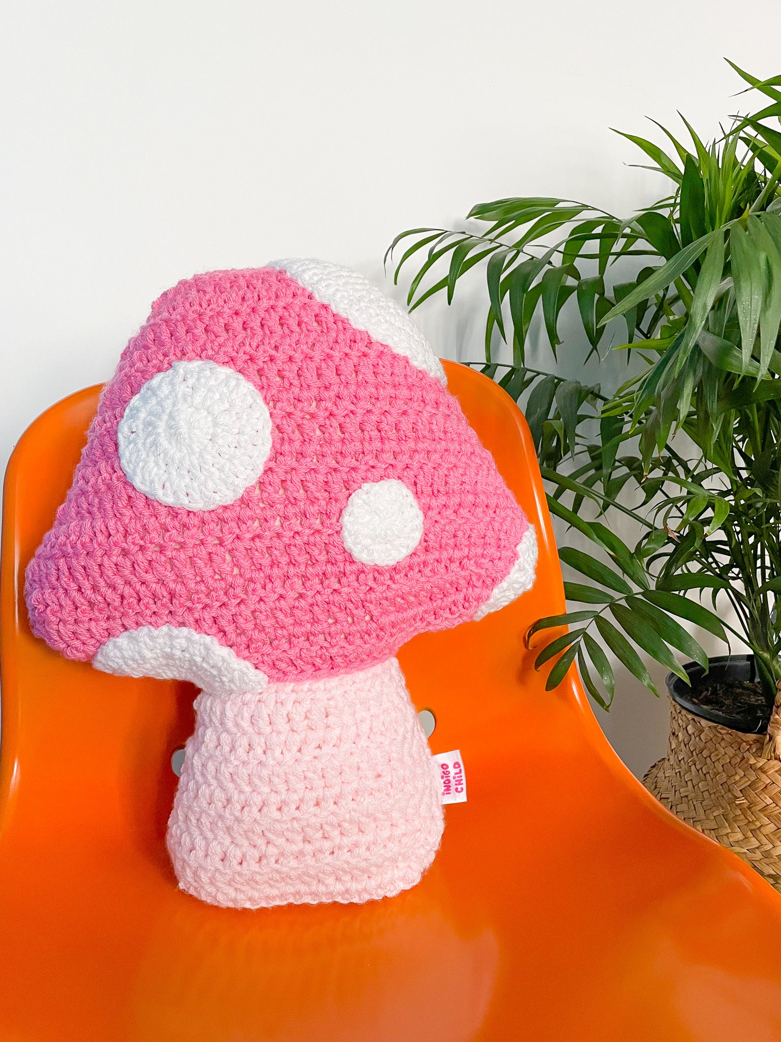 A pink mushroom shaped pillow lying on an orange desk chair with a large plant next to it.