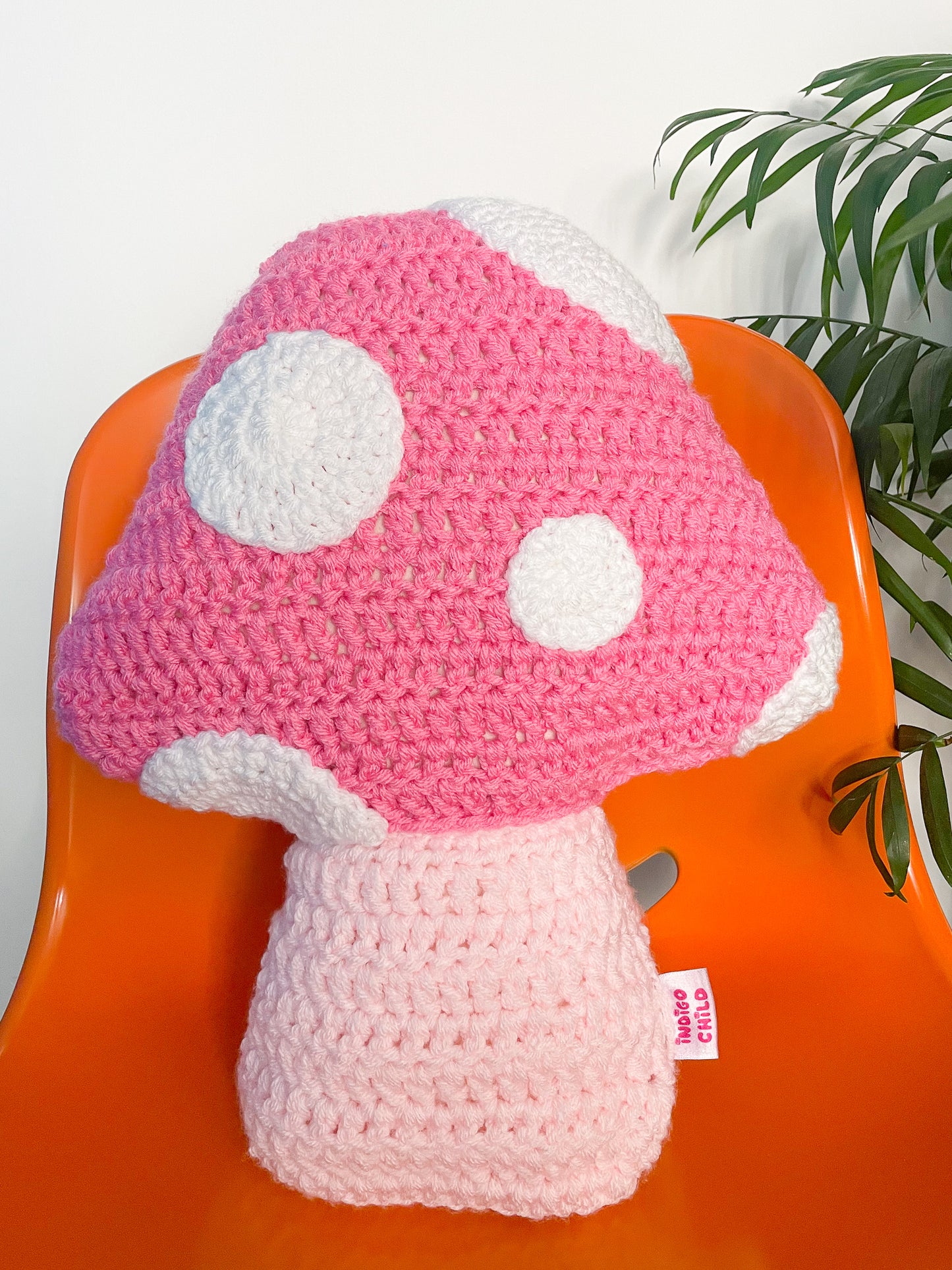 Close view of a mushroom shaped pillow in pink with white crochet appliqué dots to resemble a toadstool sat on an orange chair.