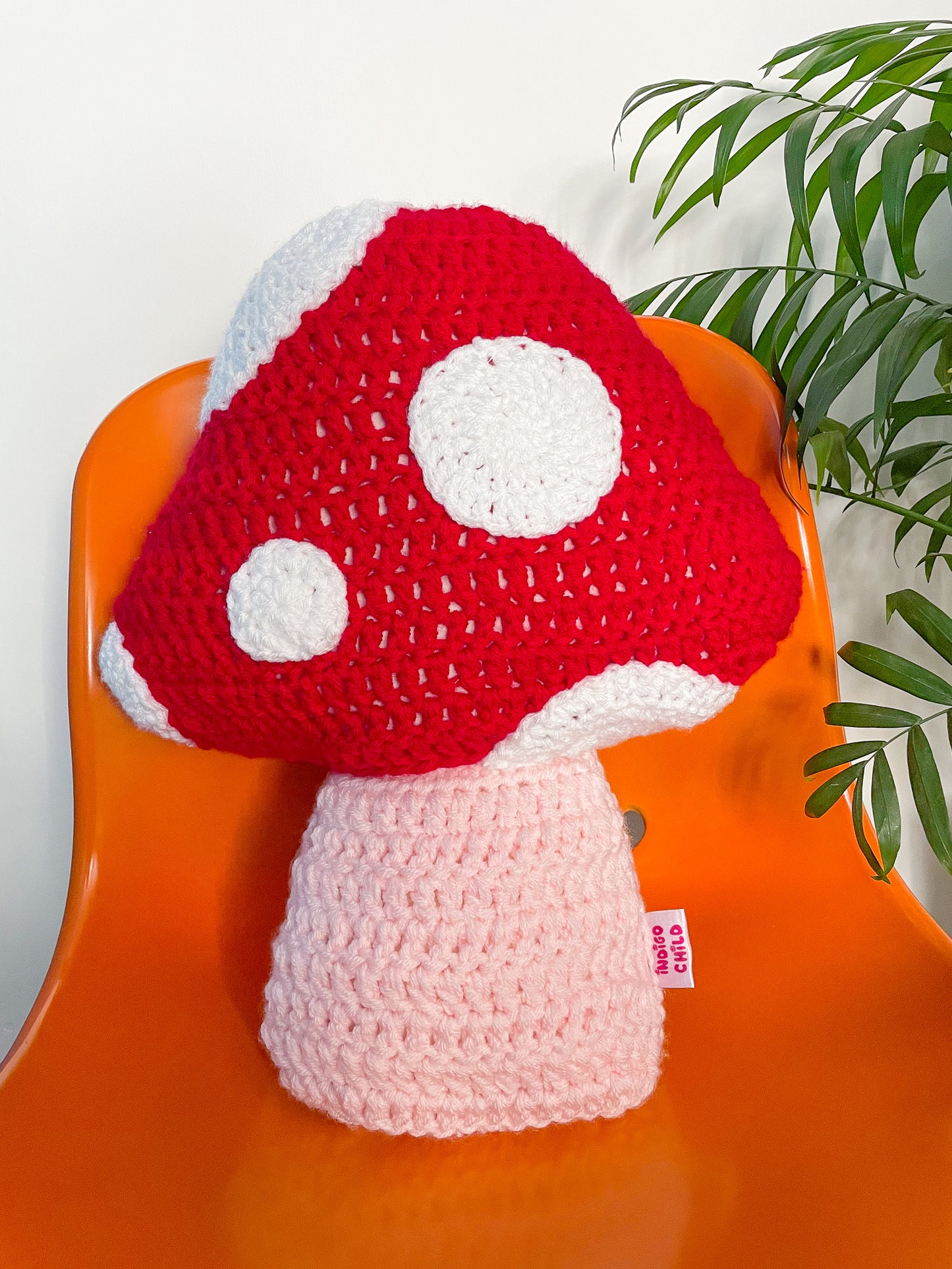 Close view of a crochet cushion shaped like a toadstool mushroom in red with white dots sewn on it sat on an orange desk chair.