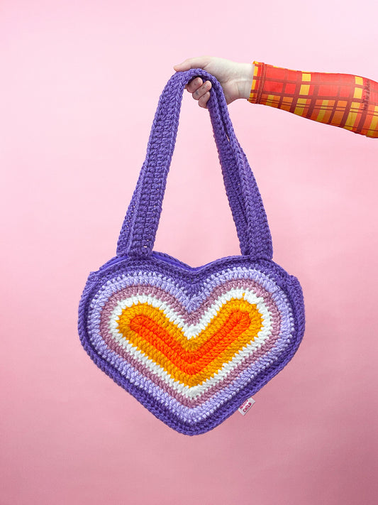 A hand holding a heart shaped bag resembling lesbian flag colours (orange, white, pink and purple) in front of a pink background.