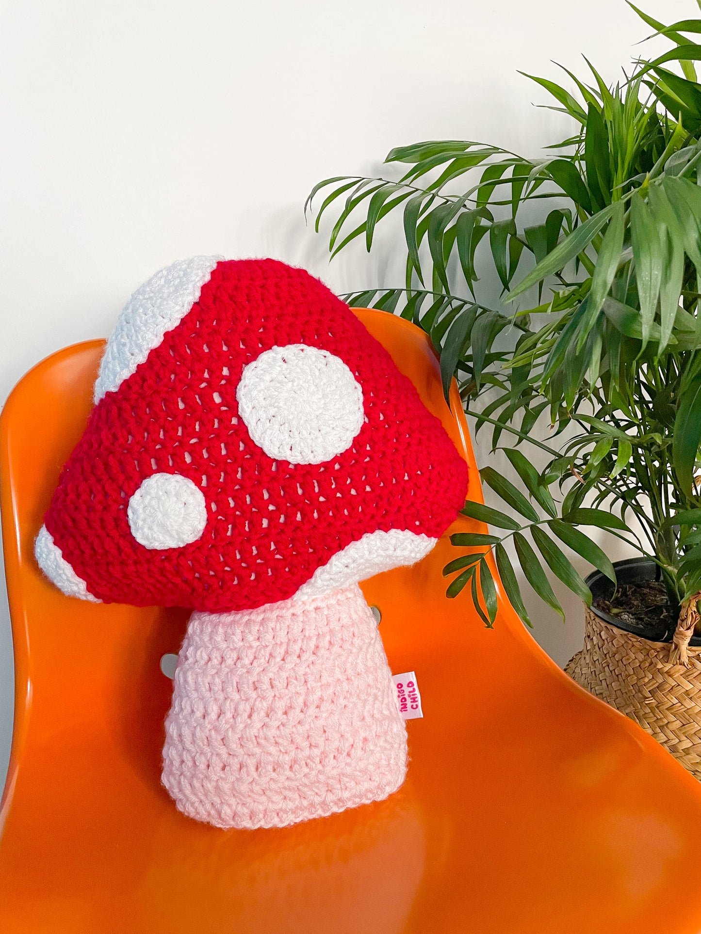 A mushroom shaped crochet pillow in red and white appliqué dots sat on an orange desk chair with a plant next to it.