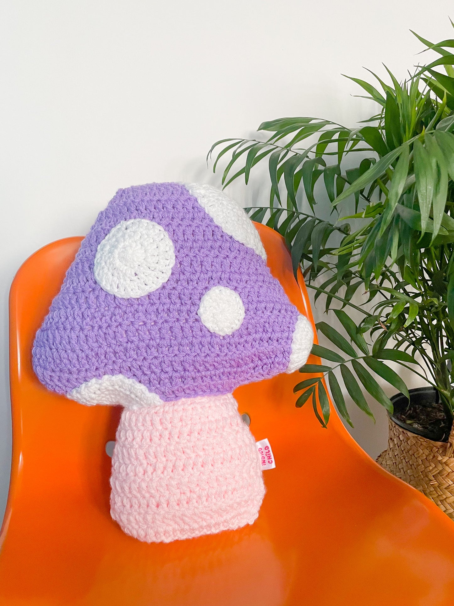 A lilac crochet cushion shaped like a toadstool mushroom with white appliqué dots on the front sat upwards on an orange desk chair next to a plant.