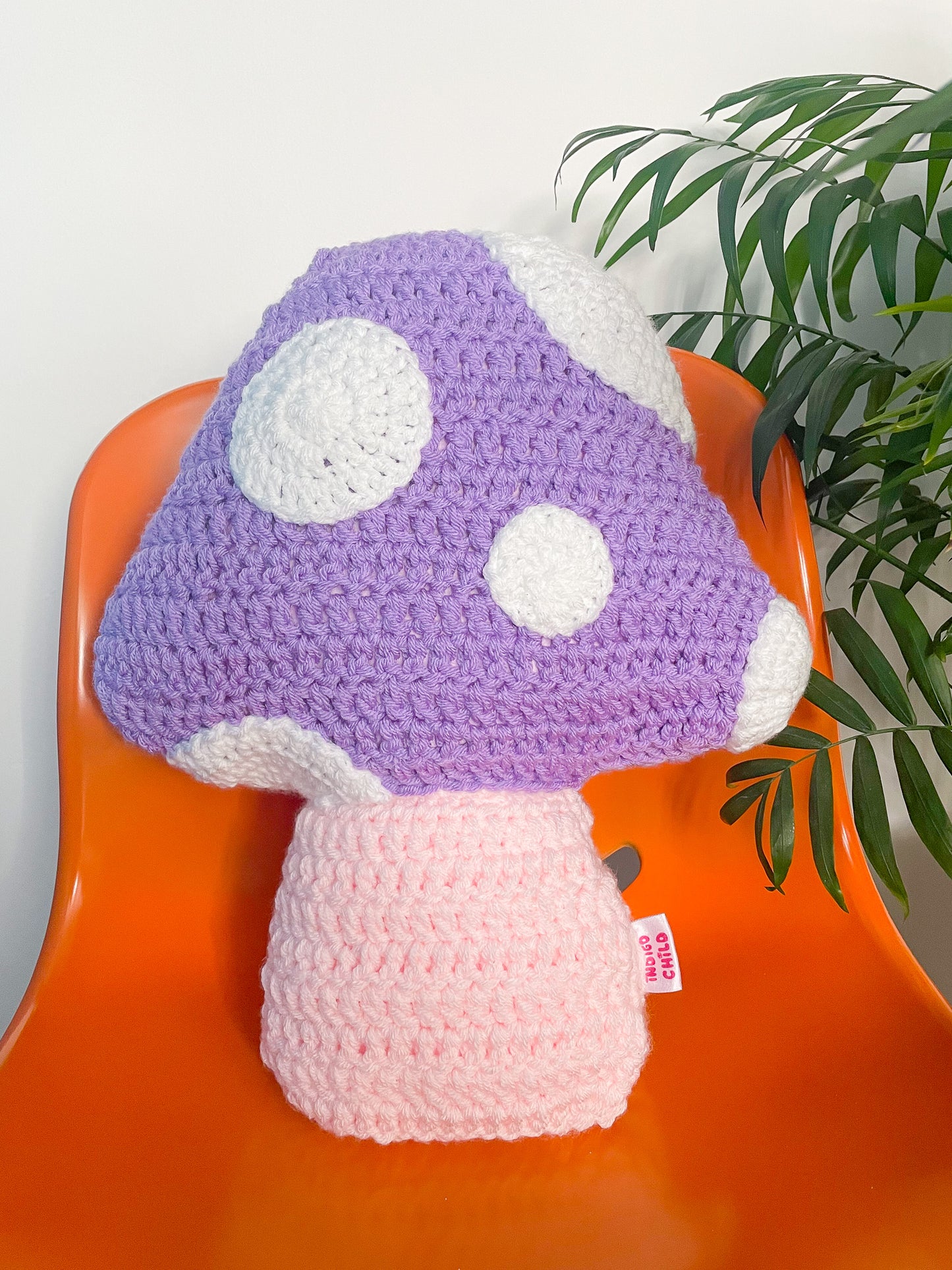 Close view of a lilac pillow shaped like a toadstool mushroom sat upright on an orange desk chair with a plant next to it.