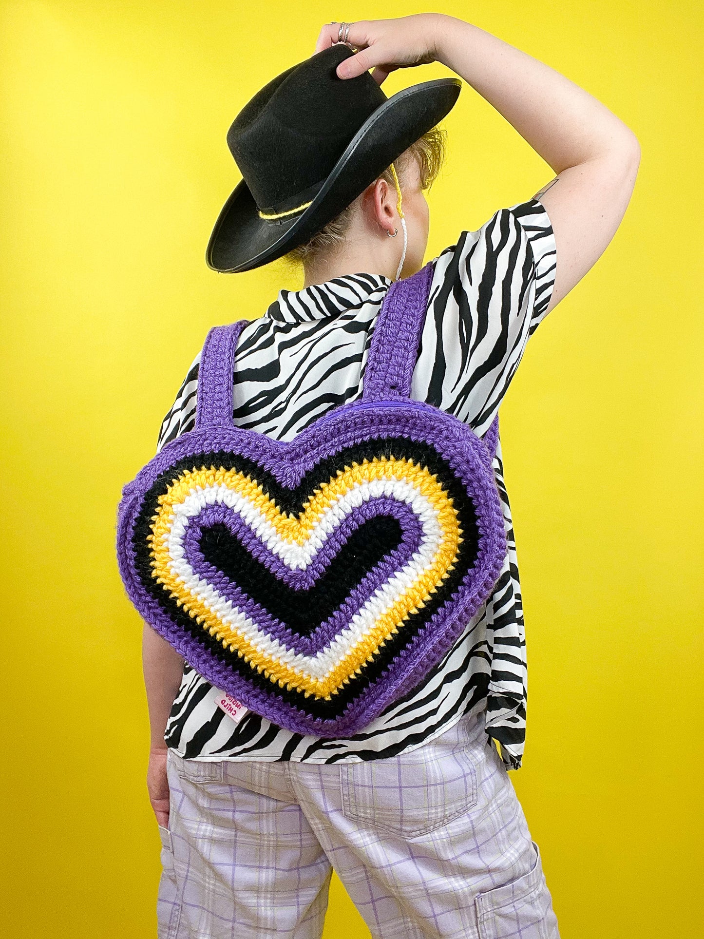 Back view of a person wearing a non binary flag bag (black, purple, white, yellow) in a heart shape with a black cowboy hat, zebra shirt and lilac jeans.