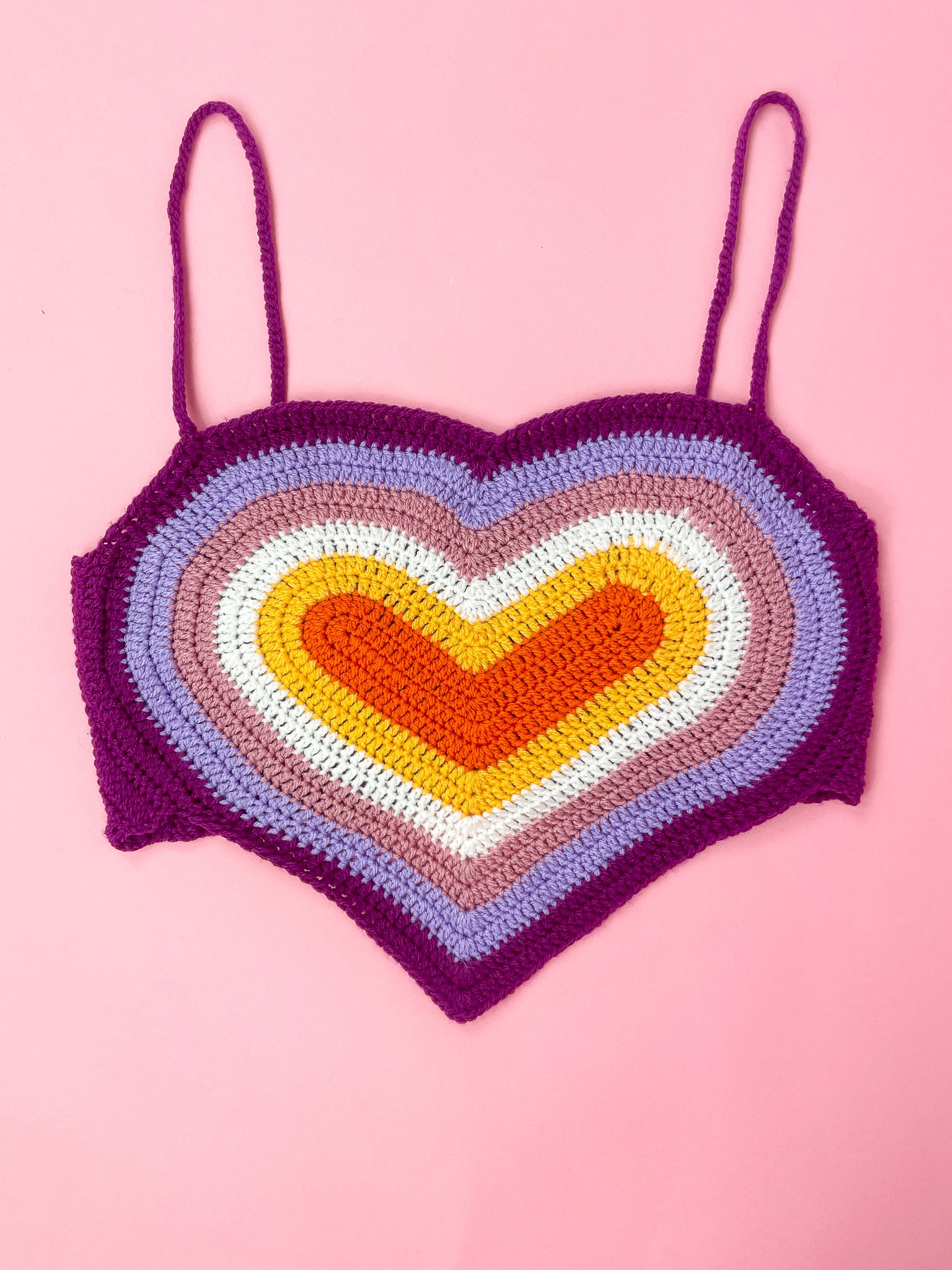 Flat lay of a crochet top shaped like a heart with a lesbian flag rainbow design (orange, pale orange, white, pale pink, lilac, purple) lying on a pale pink background.