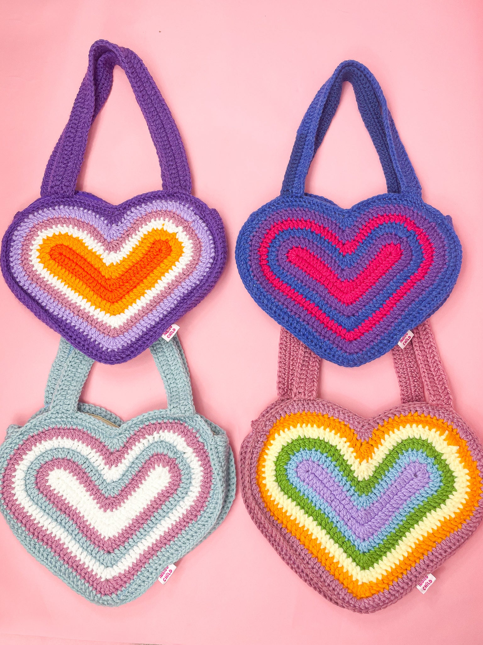 Four heart shaped bags in different pride flag colours (lesbian, bisexual, trans and pastel rainbow), lying together on a pink background.