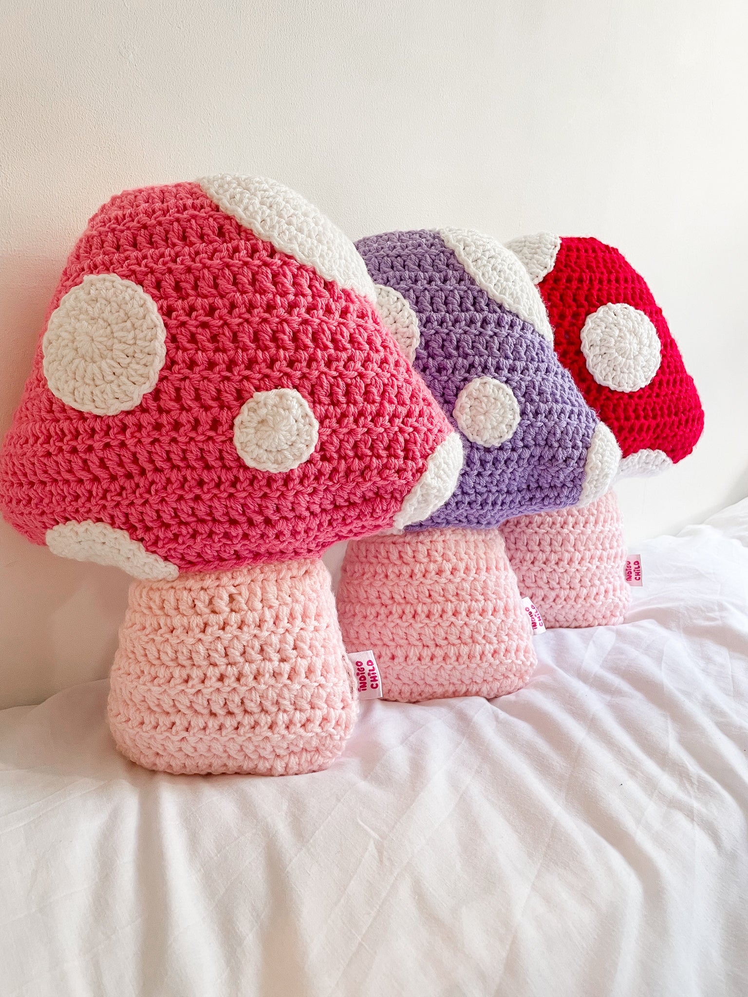 Three crochet pillows shaped like toadstool mushrooms in pink, lilac and red leaning against a white wall in a row.