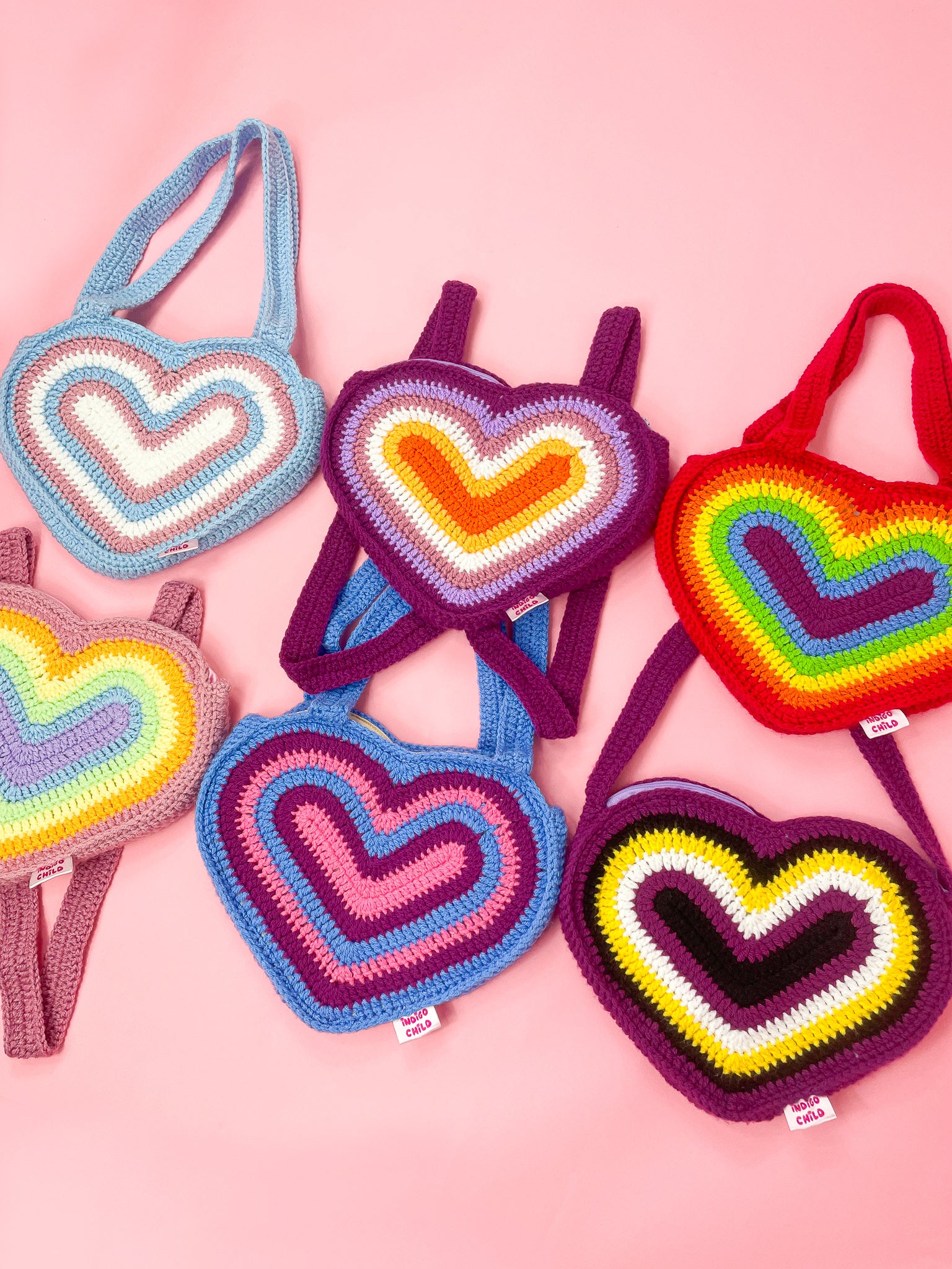 Six crochet heart bags in different pride flag colours (trans, lesbian, traditional, pastel, bisexual and non binary), lying together on a pink background.