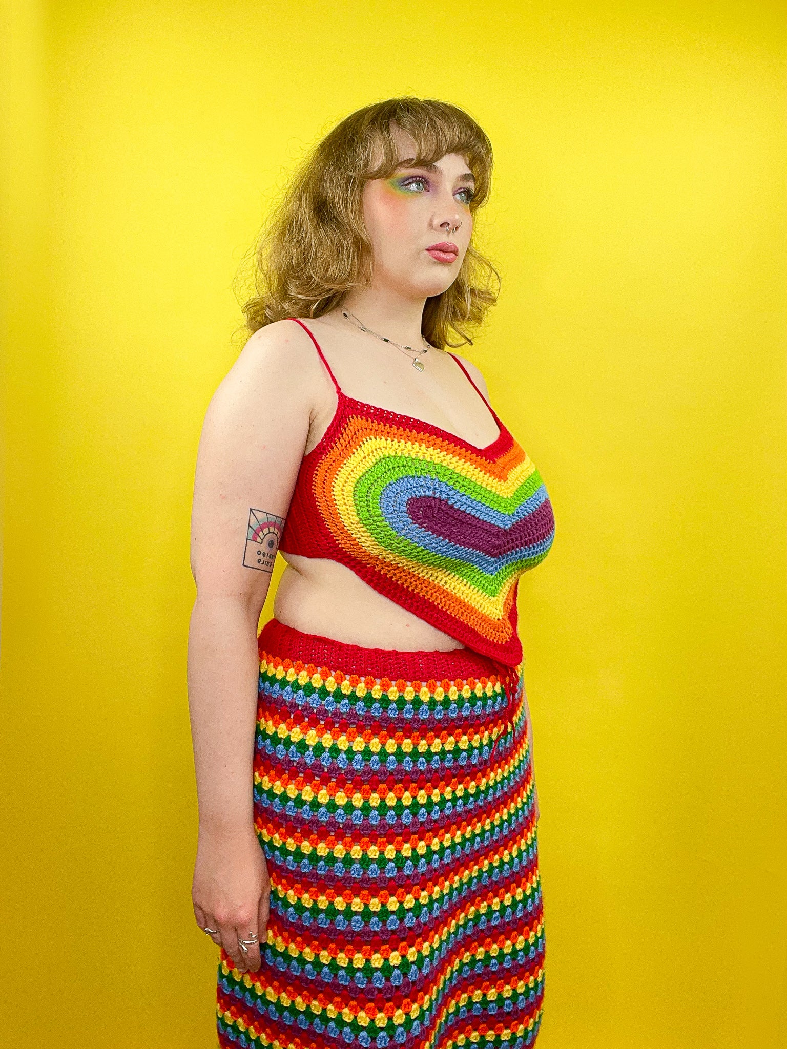 Side view of a woman wearing a bright set with a heart shaped crop top and a striped rainbow skirt.