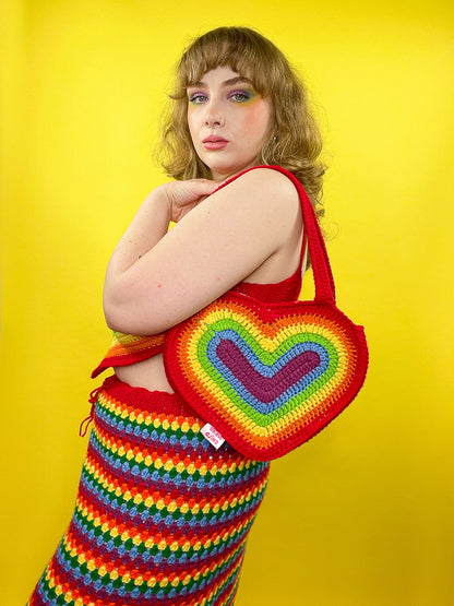 Side view of a woman wearing a rainbow heart shaped crochet bag on her shoulder with a matching vibrant top and skirt.