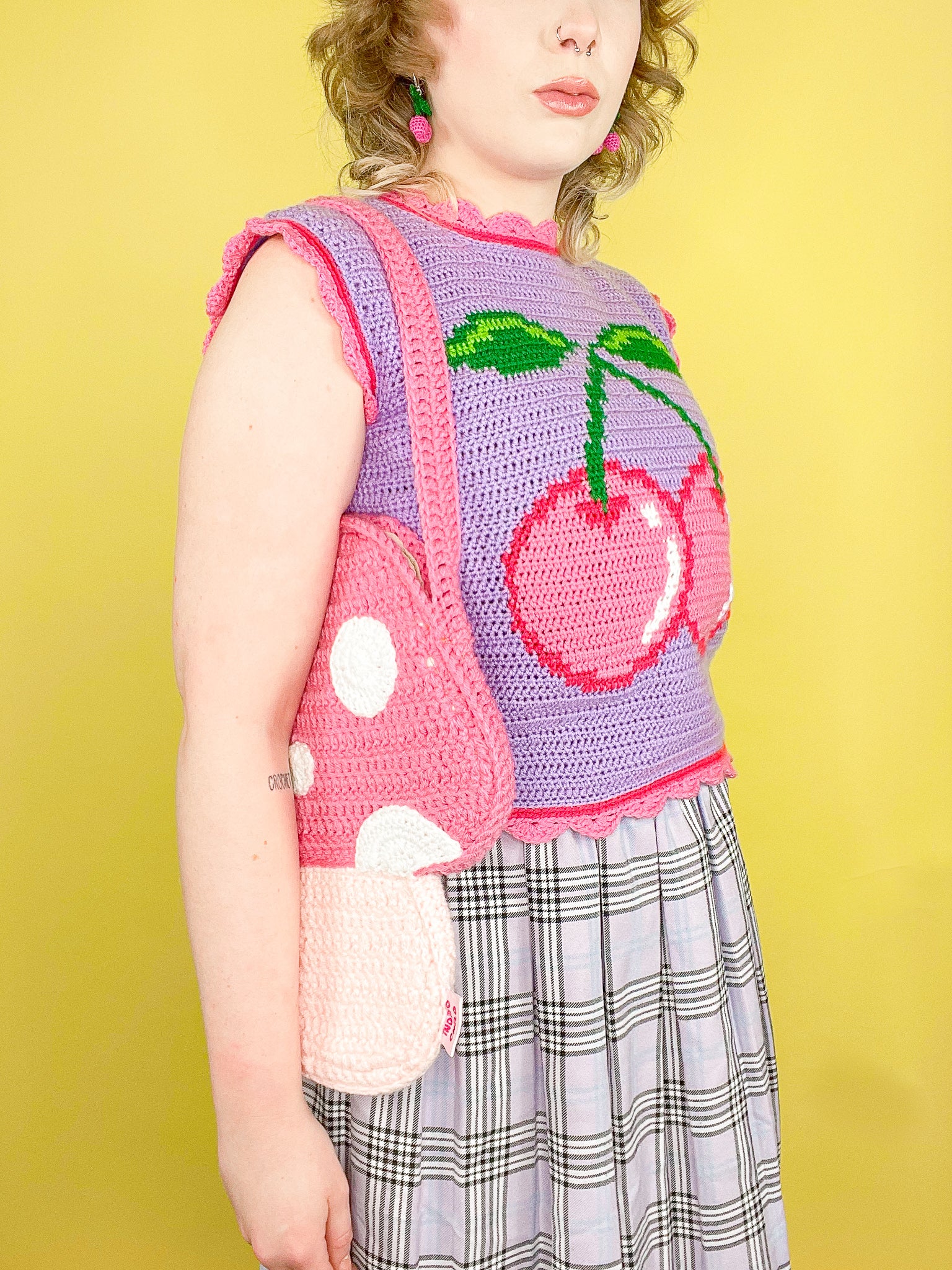 Side view of a woman wearing a toadstool mushroom bag in pink on her shoulder paired with a lilac vest that has a pink cherry on the front and a lilac kilt.